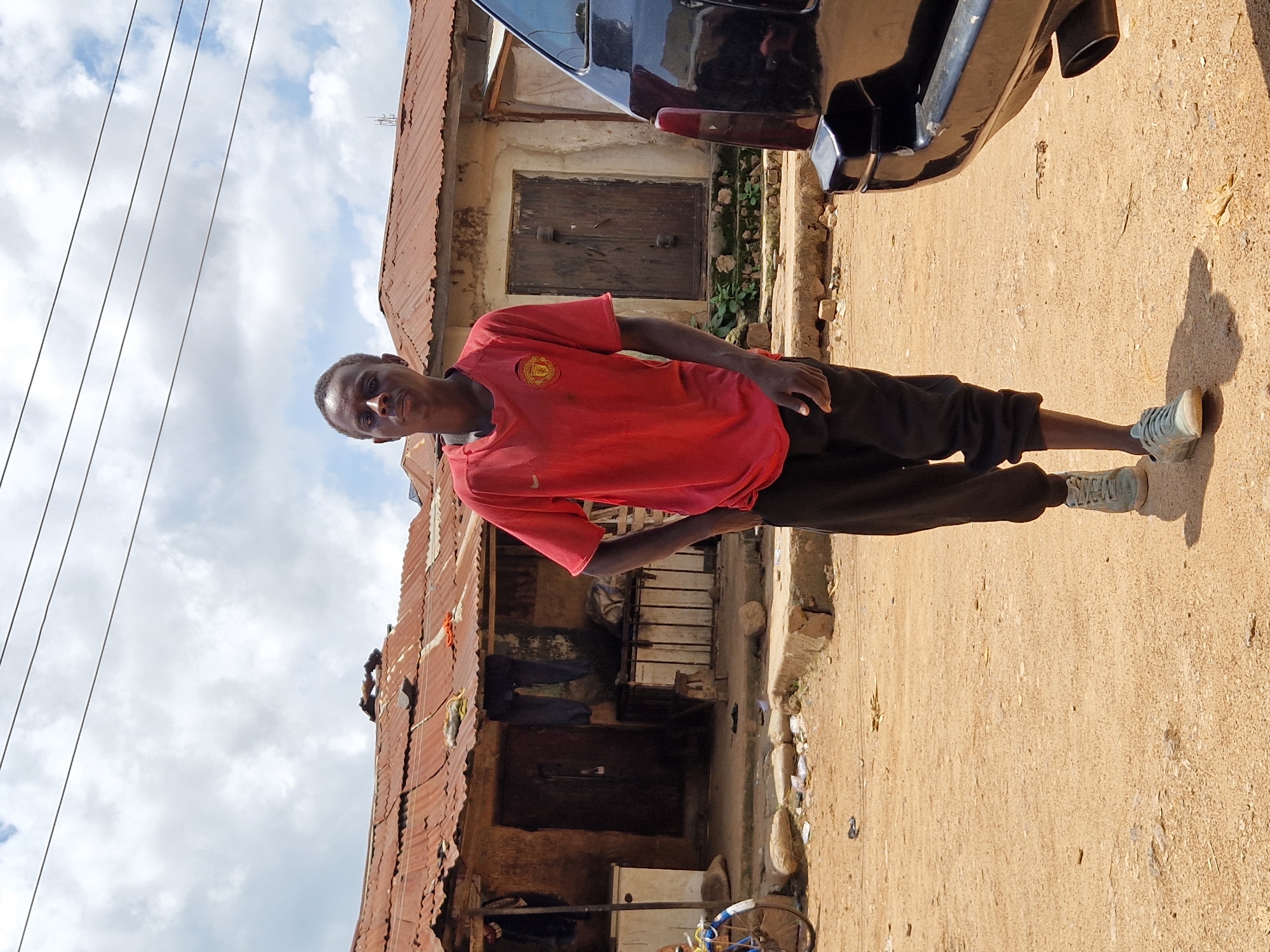 A young Black male, appearing to be in his late teens or early twenties, stands on a dirt road in Jos, Nigeria. He is wearing a red t-shirt with a crest on the left chest and dark, loose-fitting pants. His left hand is in his pocket, and his right arm is relaxed at his side. He is looking directly at the camera. He is wearing light-colored sneakers.
Behind him, a row of buildings with corrugated metal roofs, some appearing rusted and in disrepair, line the street. The buildings have wooden doors and some windows are visible. There is debris and some scattered papers on the ground in front of the buildings. A bicycle with a blue frame is partially visible on the left side of the image. A black vehicle is parked to the right of the frame, its rear bumper and taillight visible. The sky is cloudy with patches of blue. The overall scene suggests an outdoor, informal setting within a populated area.