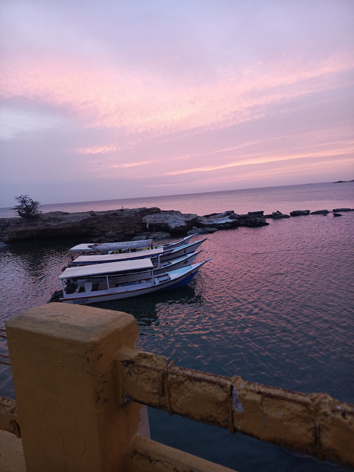 The image captures a serene sunset in Juan Griego, Venezuela. The sky is a blend of pink and purple hues, reflected in the calm waters. Three boats are docked near a rocky outcrop with sparse vegetation. A weathered yellow railing is visible in the foreground, adding depth to the scene. The boats are white and blue, one labeled "Yurath Tampapall." The horizon appears calm and undisturbed. No people are apparent in the image.