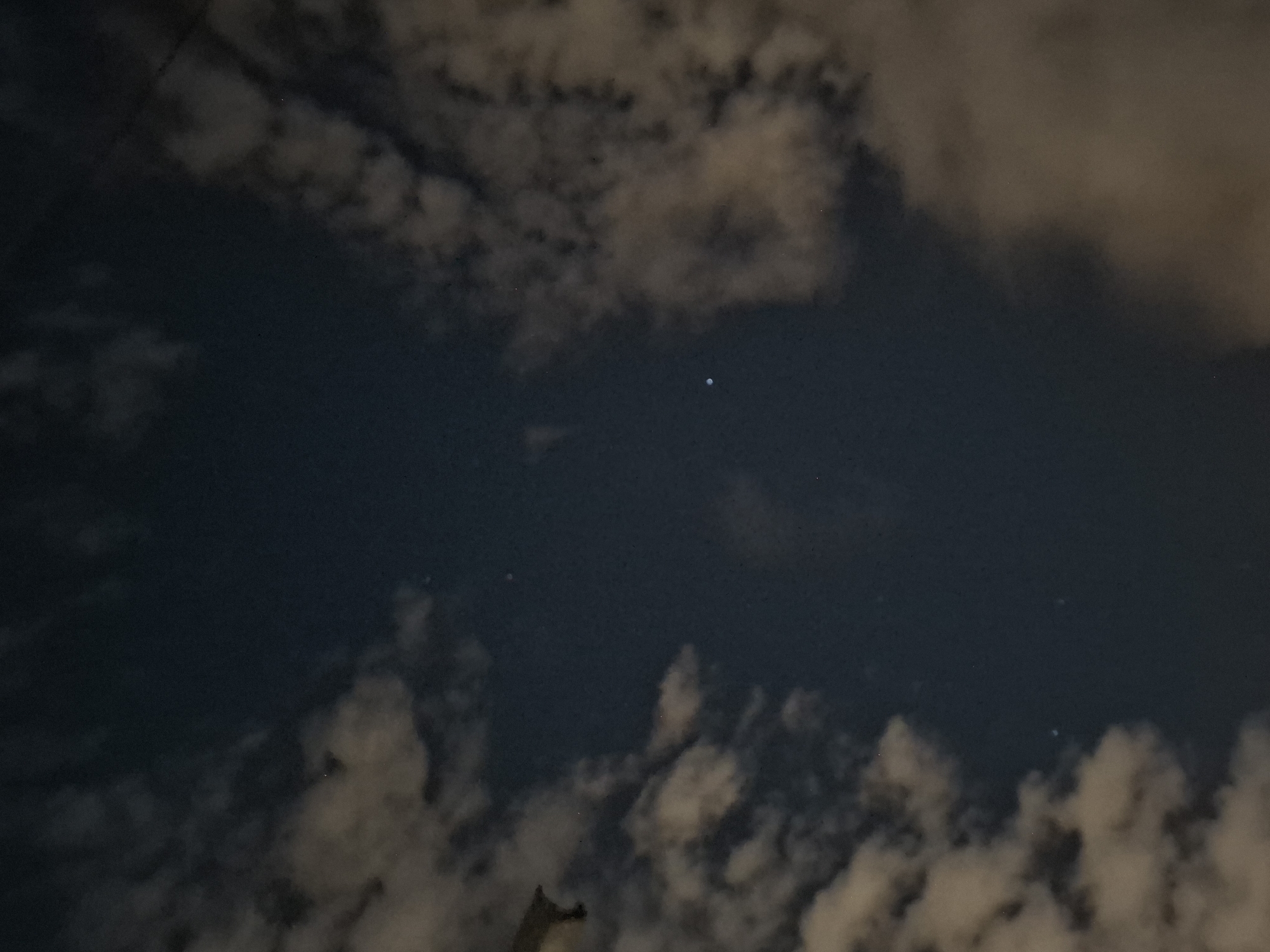The image shows a night sky with scattered clouds, illuminated by artificial light. Two bright points of light are visible, possibly stars or planets. The bottom left of the image shows a sliver of a dark object, which could be a rooftop or a building structure. The overall scene suggests an urban environment at night, with a view of the sky above Almaty, Kazakhstan. The lighting of the clouds indicates the presence of streetlights or building lights below.
