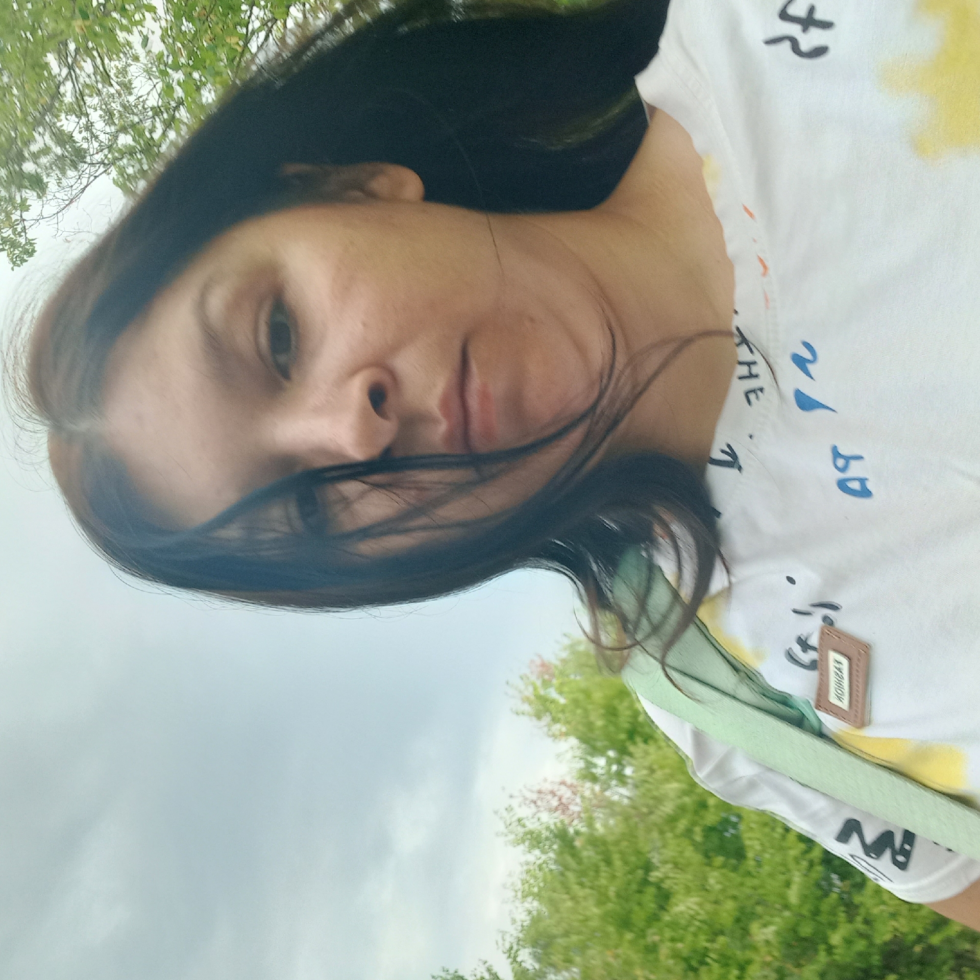 A woman with dark hair and medium-toned skin is taking a selfie outdoors. She is wearing a white t-shirt with blue and yellow abstract designs and text. She has a light green strap of a bag visible across her chest. The background consists of lush green trees and a cloudy sky. The image was taken near the village of Neopalymivka, Ukraine.