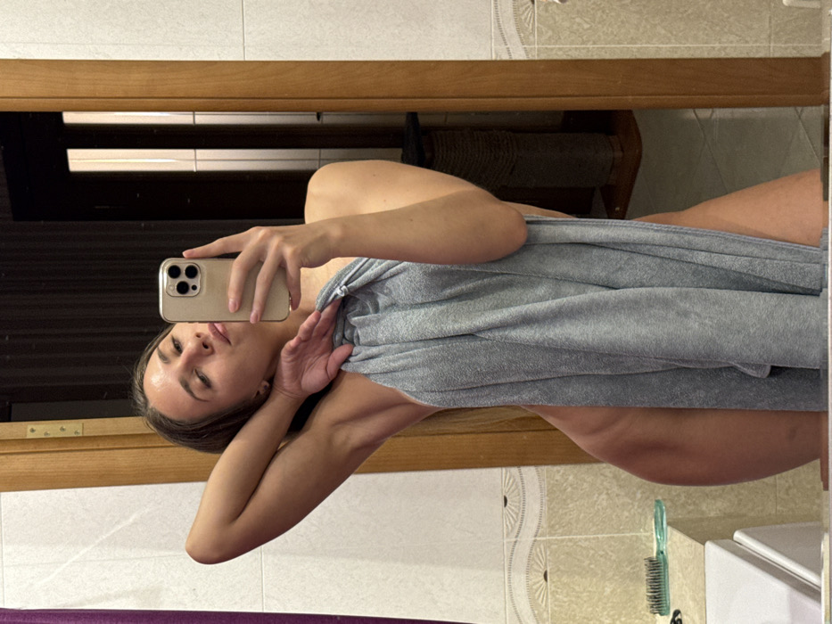 The image depicts an adult female capturing a mirror selfie within what appears to be a private residential interior, likely a bathroom or an adjacent room. The female, with dark hair, is seen from approximately mid-thigh up, wrapped in a light grey towel. Her right hand holds a light-colored smartphone, aimed at the mirror. Her left arm is raised, with her hand positioned near the back of her head. Her head is tilted slightly, and she presents a subtle pout.
The reflected environment reveals light-colored square wall tiles, surmounted by a decorative border featuring wavy lines and a fan-like motif. To the left, a portion of a white porcelain fixture, possibly a sink or bathtub, is visible, with a green hairbrush resting on its surface. A light brown wooden door frame is present on the right side of the reflection. Beyond this doorway, elements resembling a rope-wrapped cat scratching post and light-colored floor tiles are discernible. Another darker door or panel is visible further back on the left side of the reflection. The photograph contains no identifiable external landmarks or specific architectural features to confirm a precise location within the city of Saint Petersburg, Russia.