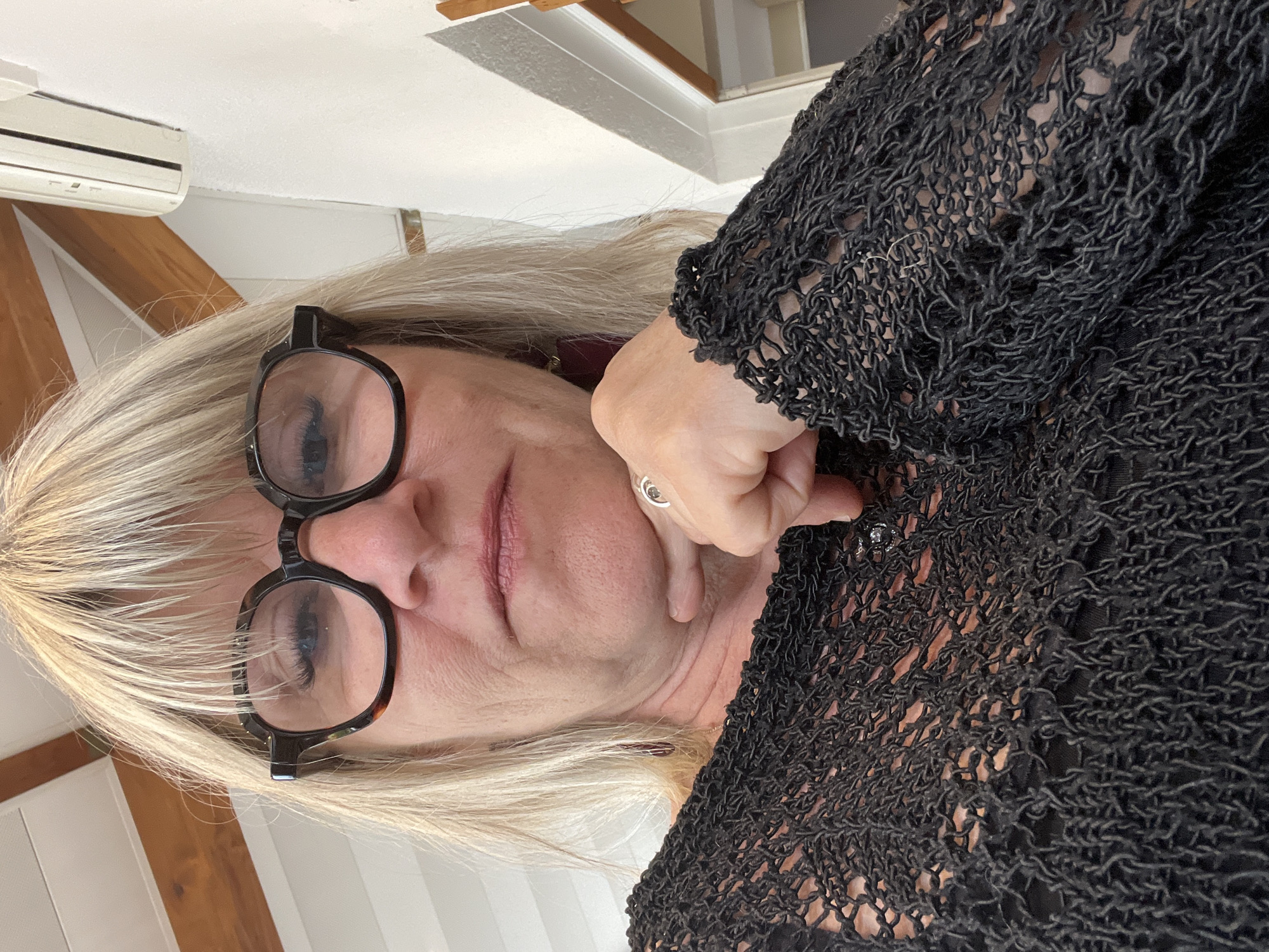 A middle-aged woman with short blonde hair, wearing glasses, is looking at the camera. She is wearing a black knitted top. The background appears to be the interior of a building, possibly with white walls and wooden beams. The location is identified as Grimaud, France. There are no explicit details to determine the precise location, so no interaction can be identified.