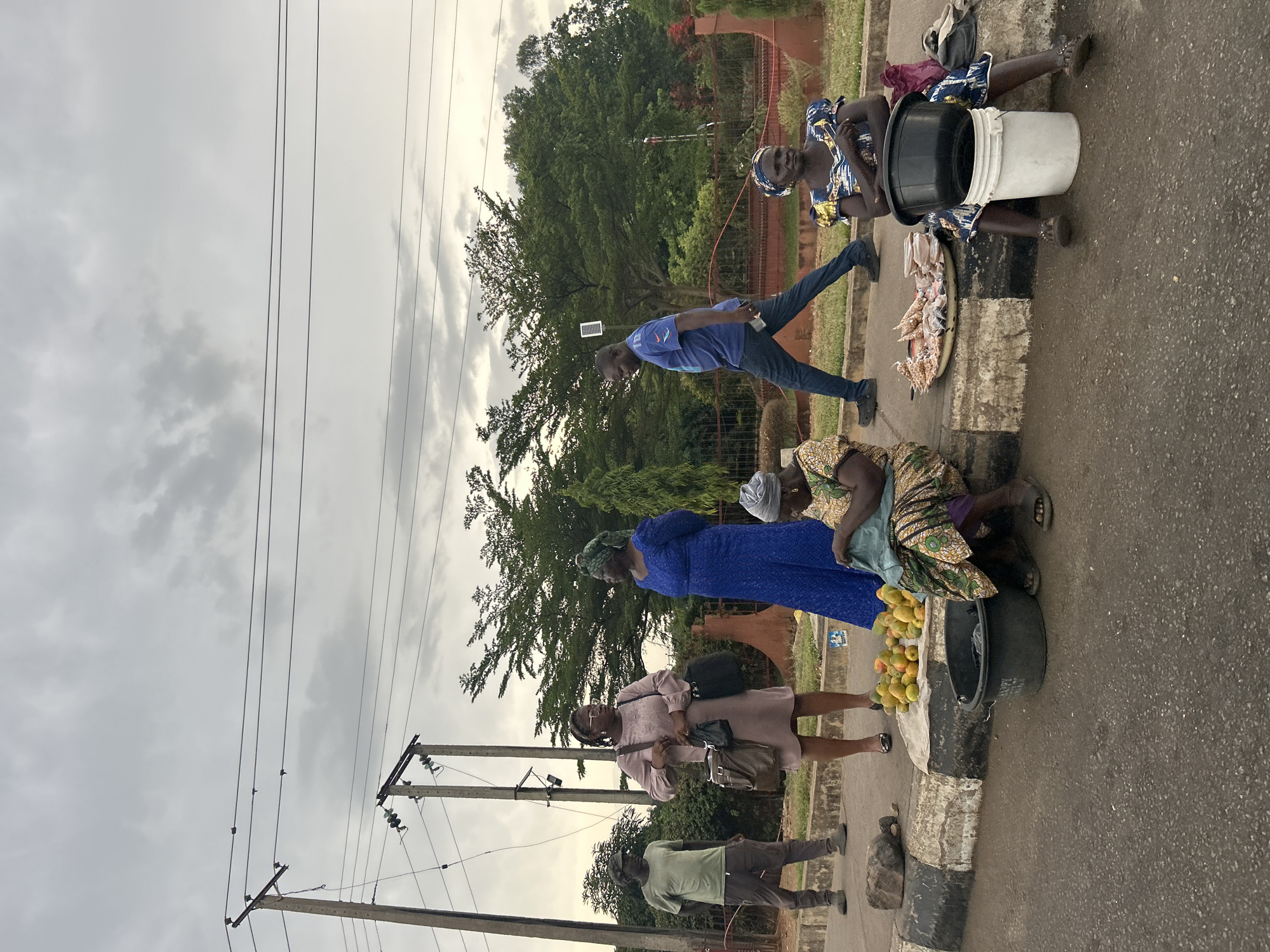 In Jos, Nigeria, a street scene unfolds on a cloudy day. Overhead, power lines stretch across the sky. Several people are present: three women appear to be vendors selling goods on the roadside; one sells mangoes and another prepared meals, possibly akara. One man stands on the road's edge, while another walks across the road, looking down. There are trees, and a building in the background.