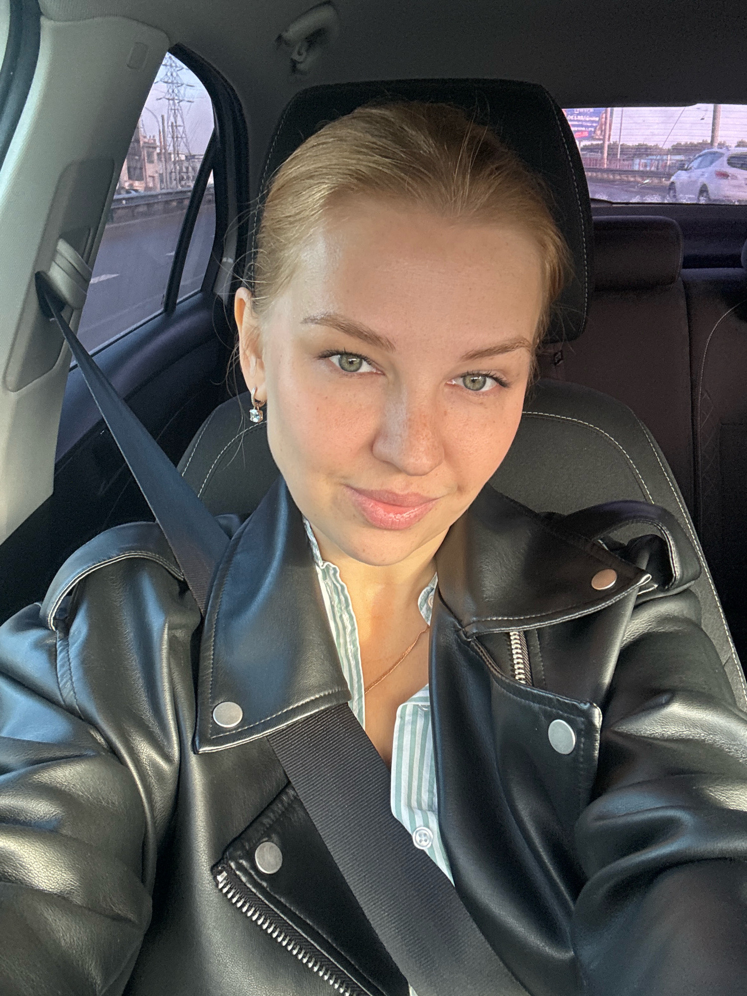 A young Caucasian woman with blonde hair pulled back and light green eyes is sitting in the driver's seat of a car. She is wearing a black leather jacket over a light green and white striped button-up shirt and a delicate gold necklace. A black seatbelt is fastened across her chest. She is looking at the camera with a slight smile. Freckles are visible on her cheeks and nose. The interior of the car, including the black headrest and seat, is visible. Through the side window, power lines and industrial buildings are seen, suggesting an urban or industrial environment. The scene is consistent with a selfie taken while driving, possibly in the vicinity of Rusanovo, Russia, given the context.