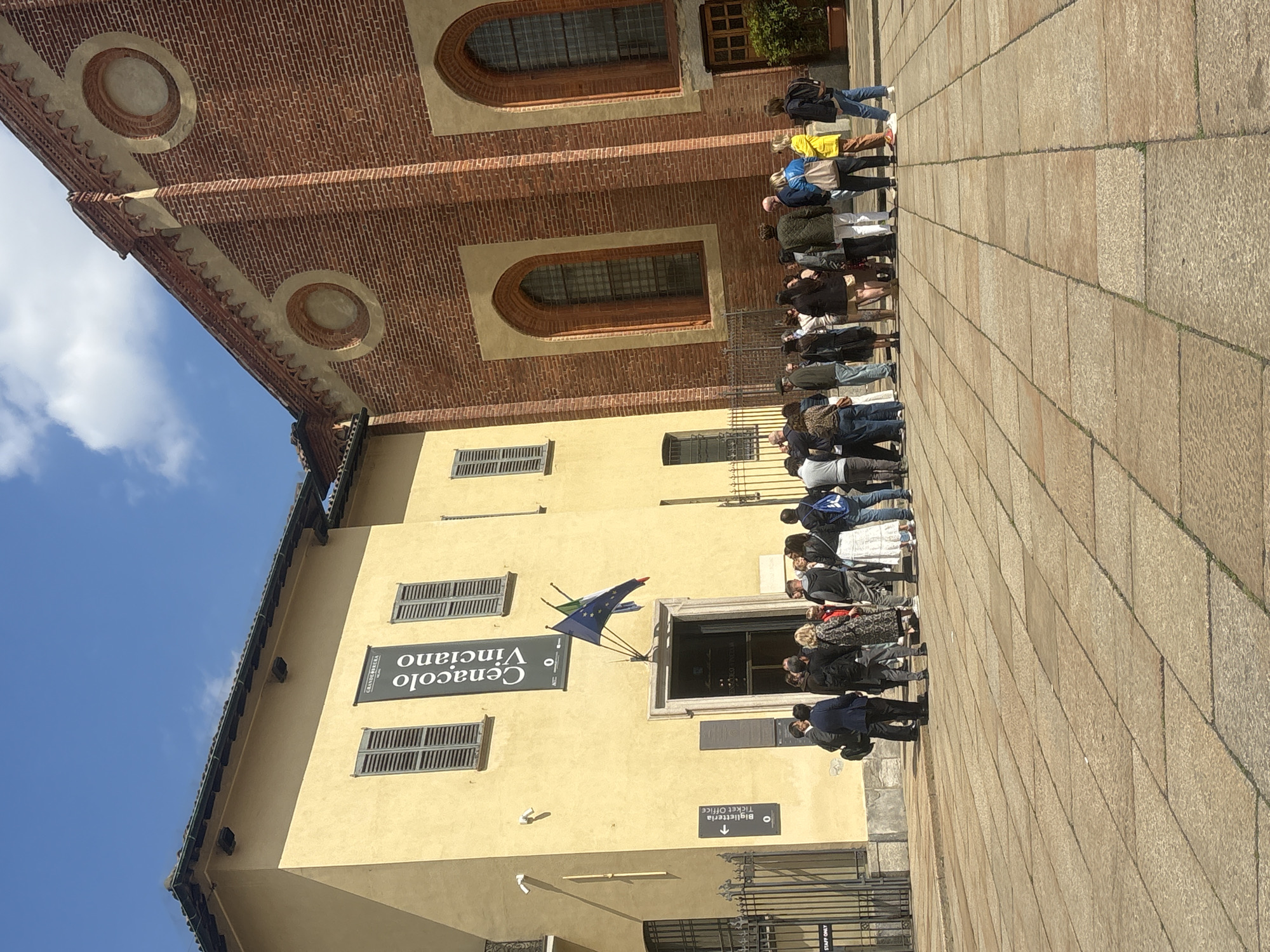 The image, rotated 90 degrees clockwise, captures an outdoor scene featuring a queue of people in front of historical buildings under a clear blue sky with some scattered clouds. The foreground shows a wide, paved area made of large, light-colored stone slabs.
On the left, a tall, yellow building dominates, characterized by rectangular windows fitted with dark, slatted shutters. Near its main entrance, two flags are displayed: the Italian flag and the flag of the European Union. A prominent dark sign attached to the yellow building reads "Cenacolo Vinciano" (referring to Leonardo da Vinci's The Last Supper) with an arrow pointing right. Below this, "Granato Plazza" is also visible. Another sign near a door indicates "Biglietteria Ticket Office" with a downward arrow. Further down, a partially visible sign reads "STAFF ONLY".
Behind the yellow building, a larger, brick-red building rises, featuring elaborate brickwork, arched windows, and circular decorative elements on its facade.
A long line of people, dressed in various casual clothing, extends from the entrance of the yellow building, along the facade, and seemingly continues towards the brick-red structure. They appear to be visitors or tourists patiently waiting to enter, likely to view the famous "Cenacolo Vinciano," as indicated by the sign. The scene is brightly lit by sunlight, suggesting daytime. The architectural style and visible text confirm the location as a historical site in Milan, Italy.