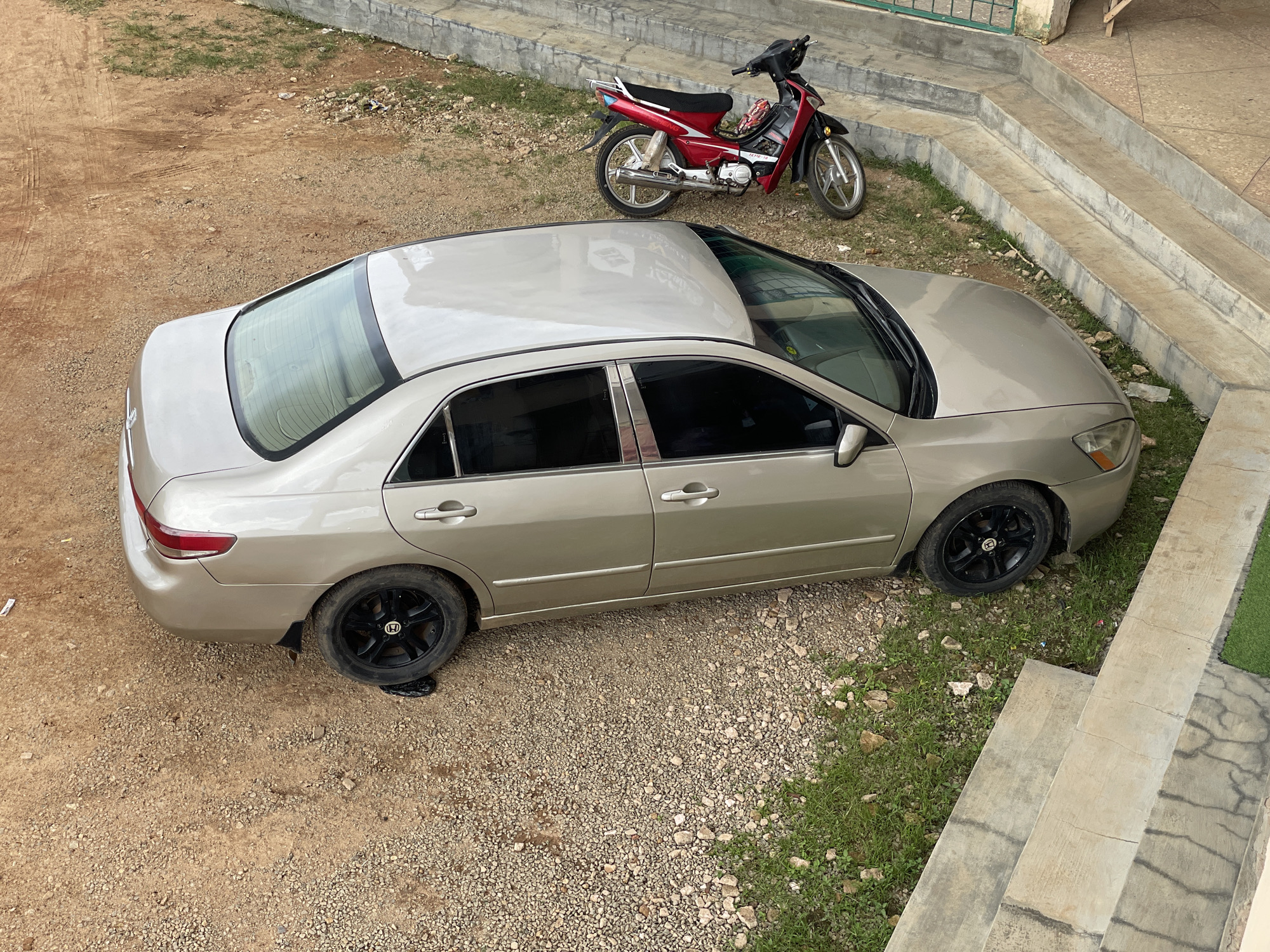In Jos, Nigeria, a gold sedan is parked on a dirt and gravel surface. The car's rear driver-side tire is flat and resting on a dark object, possibly a piece of plastic or fabric. To the left of the car, a red and black motorcycle is parked. In the background, there are concrete steps leading up to a building, with a green metal railing visible at the top. The environment appears to be an outdoor area, likely a residential or commercial space. The scene is static, with no people or apparent activity.