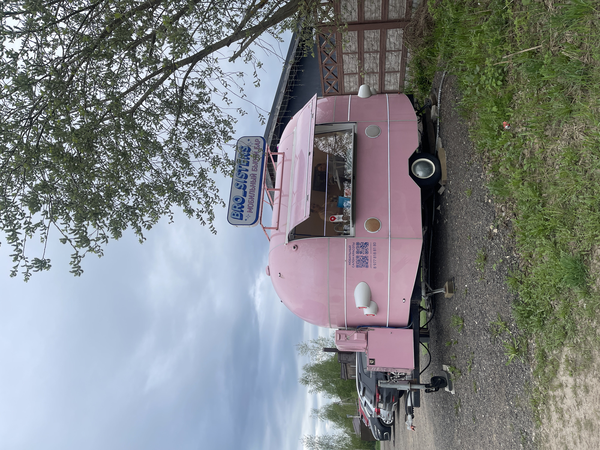 The image shows a pink "BRO SISTERS" mobile beauty salon in the city of Хлябово, Russia. The salon is a small trailer parked on a gravel path next to a patch of grass. A sign with the salon's name, "BRO SISTERS," is mounted on top. A tree with light green foliage is visible in the upper left corner of the image. Cars are parked behind the trailer. A building is behind the trailer, with a stone-clad facade. The sky is overcast.