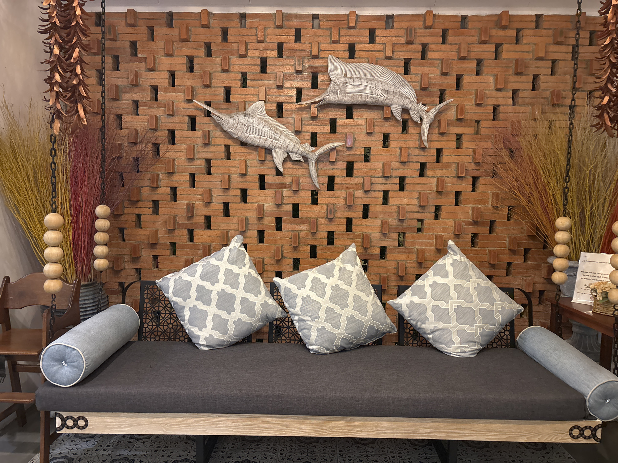 This is a photograph of a decorative wall featuring two large fish sculptures mounted on a textured brick wall. In front of the wall is a dark grey sofa with three patterned square pillows and two cylindrical bolsters. To the left of the sofa, a wooden chair and a large vase with dried decorative reeds are visible. To the right, a table with a sign and some decorative items can be seen. The overall setting appears to be an indoor space, possibly a restaurant or lounge, located in Chalong, Thailand.