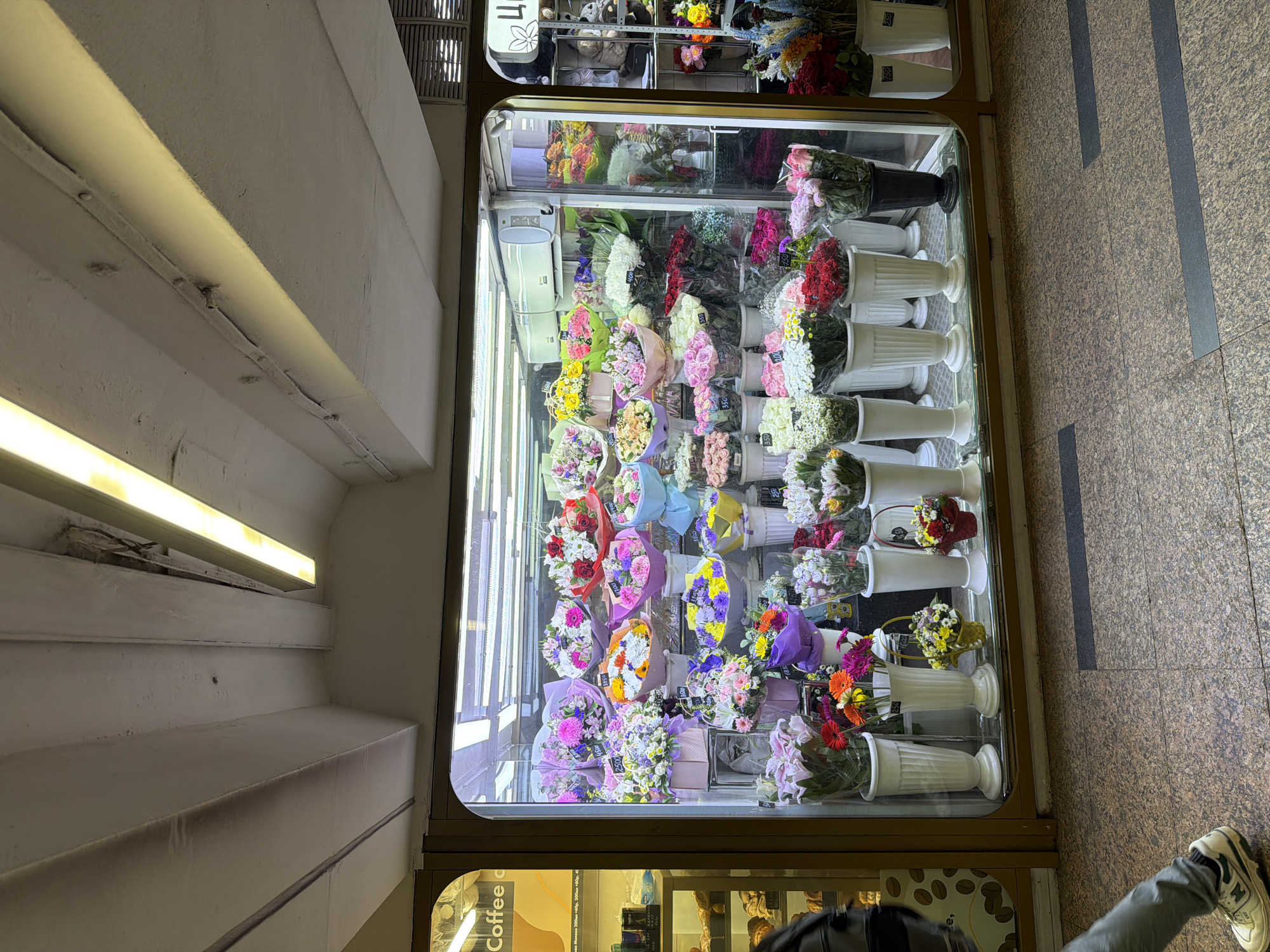 A retail display case filled with bouquets of flowers sits in the city of Unknown, Unknown, behind a glass screen, illuminated by a strip of lights from above. The flowers, wrapped in various colored papers, are arranged in white vases. The lighting inside the display case is bright, enhancing the colors of the flowers. Below the flower display is another section of the display case showing a cafe or bakery, likely selling pastries and coffee. The environment is urban, indicated by the pedestrian traffic with a person's leg partially visible in the bottom right corner. The floor is tiled. The display case is situated within a building or structure, with overhead architectural details visible on the left side of the frame.