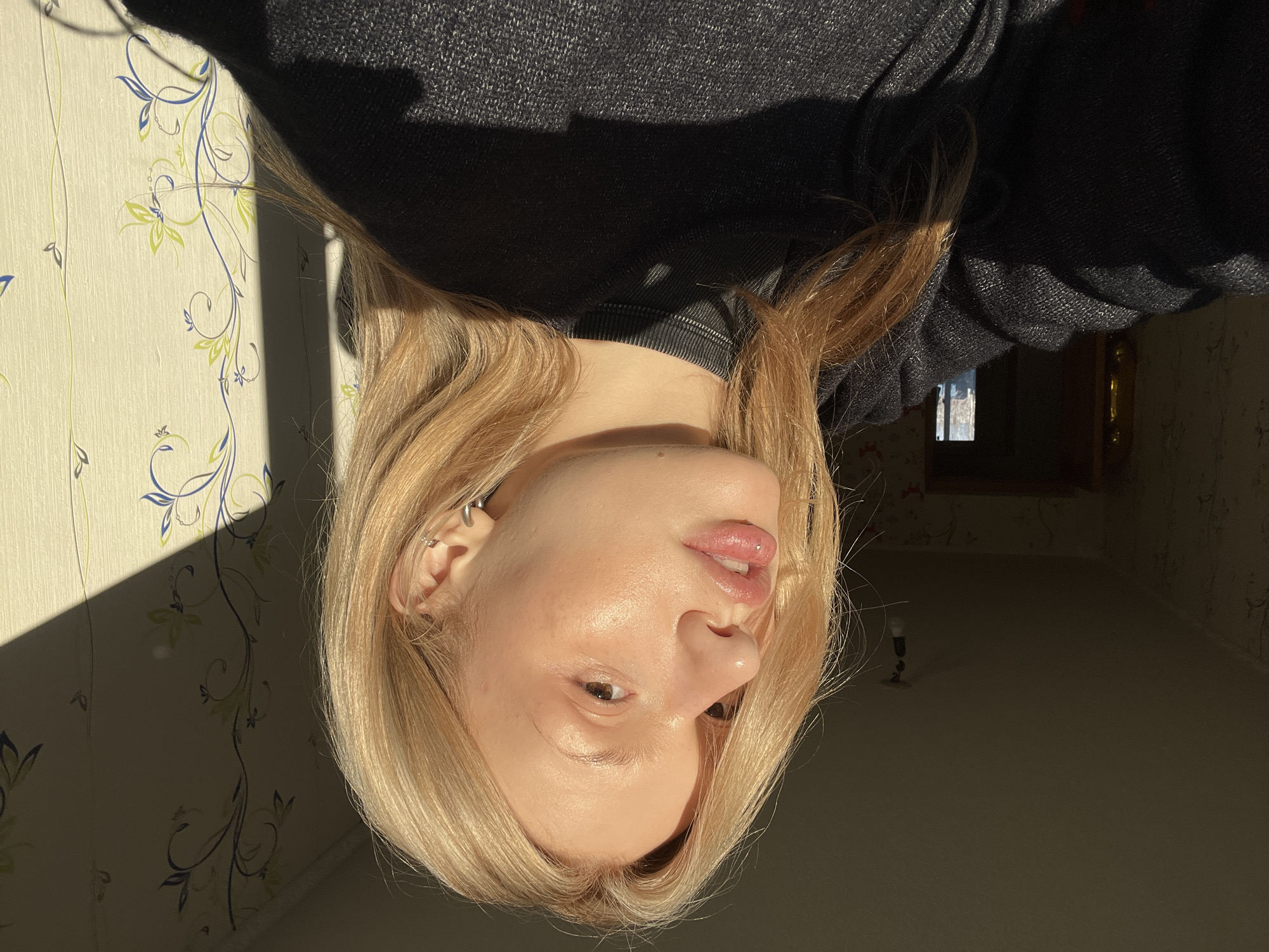 The image depicts a close-up indoor portrait of a young Caucasian female, likely engaged in taking a selfie, illuminated by strong, natural sunlight. The subject has long, straight blonde hair and fair skin, with visible freckles across her nose and cheeks. She is looking towards the right, with her lips slightly parted, revealing a silver labret piercing on her lower lip. She wears a small silver hoop earring in her lower left earlobe and a silver stud or barbell higher on the helix of the same ear. Her attire consists of a dark-colored, possibly black or dark grey, ribbed sweater or top.
Direct sunlight from the right casts bright light on the right side of her face and the adjacent wall, while the left side of her face and body, and the left portion of the room, remain in deep shadow. The background reveals a light-colored wall with a repeating pattern of blue and light green floral or vine motifs. In the shadowed area to the left, a dark doorway or archway is visible, through which a brighter, indistinct background, possibly an exterior view or another brightly lit room, can be seen. A bare light bulb hangs from the ceiling in the upper left background. The room's ceiling appears uniform and dark due to the shadows. This scene is located in a residential setting within the city of Chaikovsky, Russia.