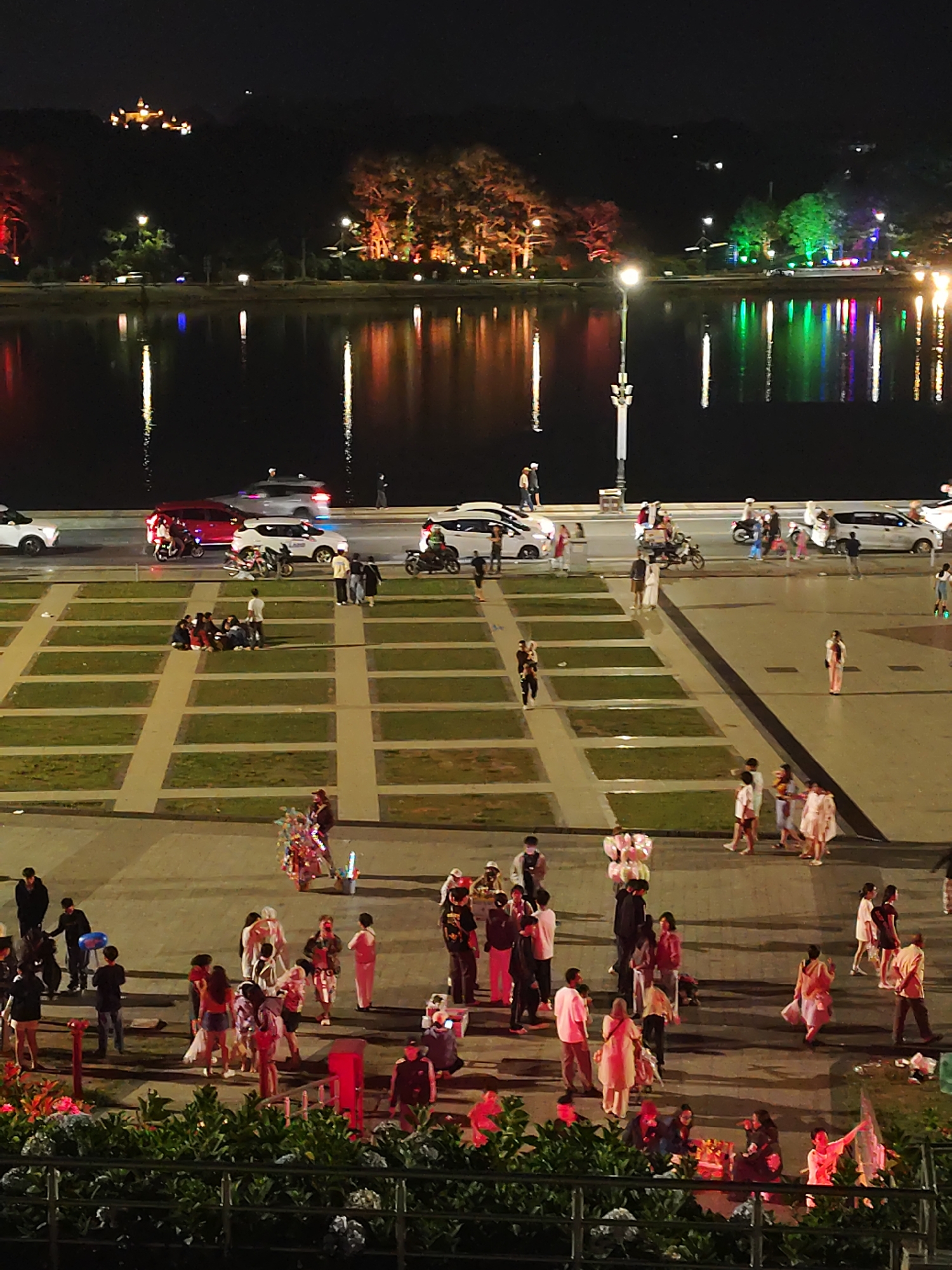 This is a nighttime aerial shot of Xuan Huong Lake in Da Lat, Vietnam. The lake's dark water reflects the vibrant lights from the trees and buildings lining its shore, creating a colorful spectacle. Several cars and motorcycles are parked along the lakeside road, with people milling about on the pedestrian walkway. In the foreground, a large plaza with checkered grassy sections is visible, also populated by groups of people. Some individuals are seated on the grass, while others stand and walk. Further down, a street vendor is seen with a cart displaying colorful items. The overall scene depicts a lively evening atmosphere in a popular urban recreational area.