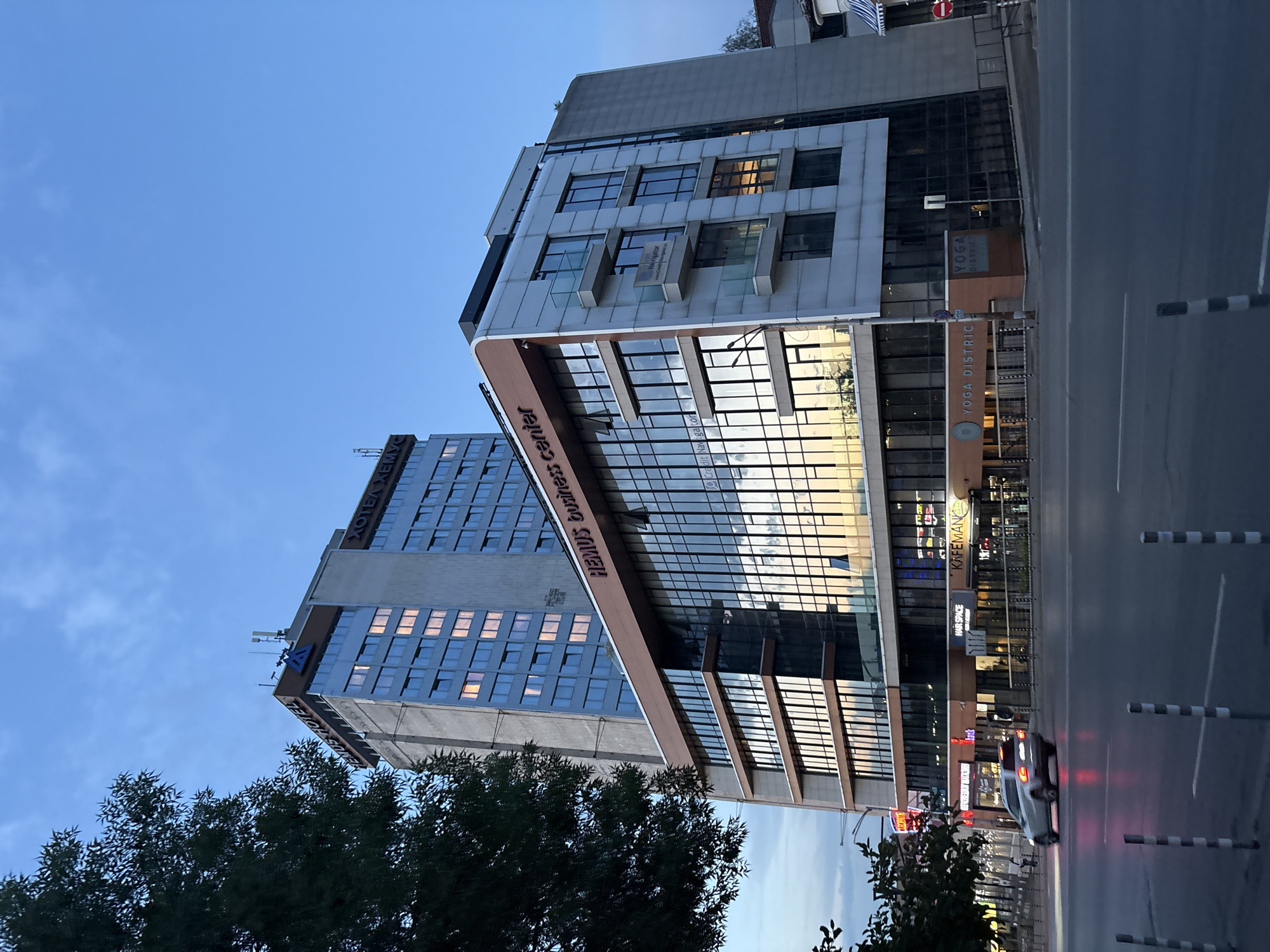 The image shows the "Hemus Hotel" and the "Hemus Business Center" in Sofia, Bulgaria, on a cloudy day at dusk. The Hemus Hotel is a tall, older building with a blue glass facade and illuminated windows, topped with a sign displaying the hotel's name and a blue logo. Adjacent to it is the Hemus Business Center, a modern, multi-story building with a glass facade that reflects the sky and surrounding environment. Various establishments, including "Hair Space," "Kafemania," and "Yoga District," are visible on the ground floor of the business center. A silver SUV is driving on the wet street in front of the buildings, its headlights on, and its taillights are visible. The street is illuminated by streetlights and the lights from the buildings. The sky is a dusky blue with some clouds, and a large tree with dark green foliage is partially visible on the left side of the frame. The scene suggests a city environment, likely an urban area with commercial and residential buildings.
