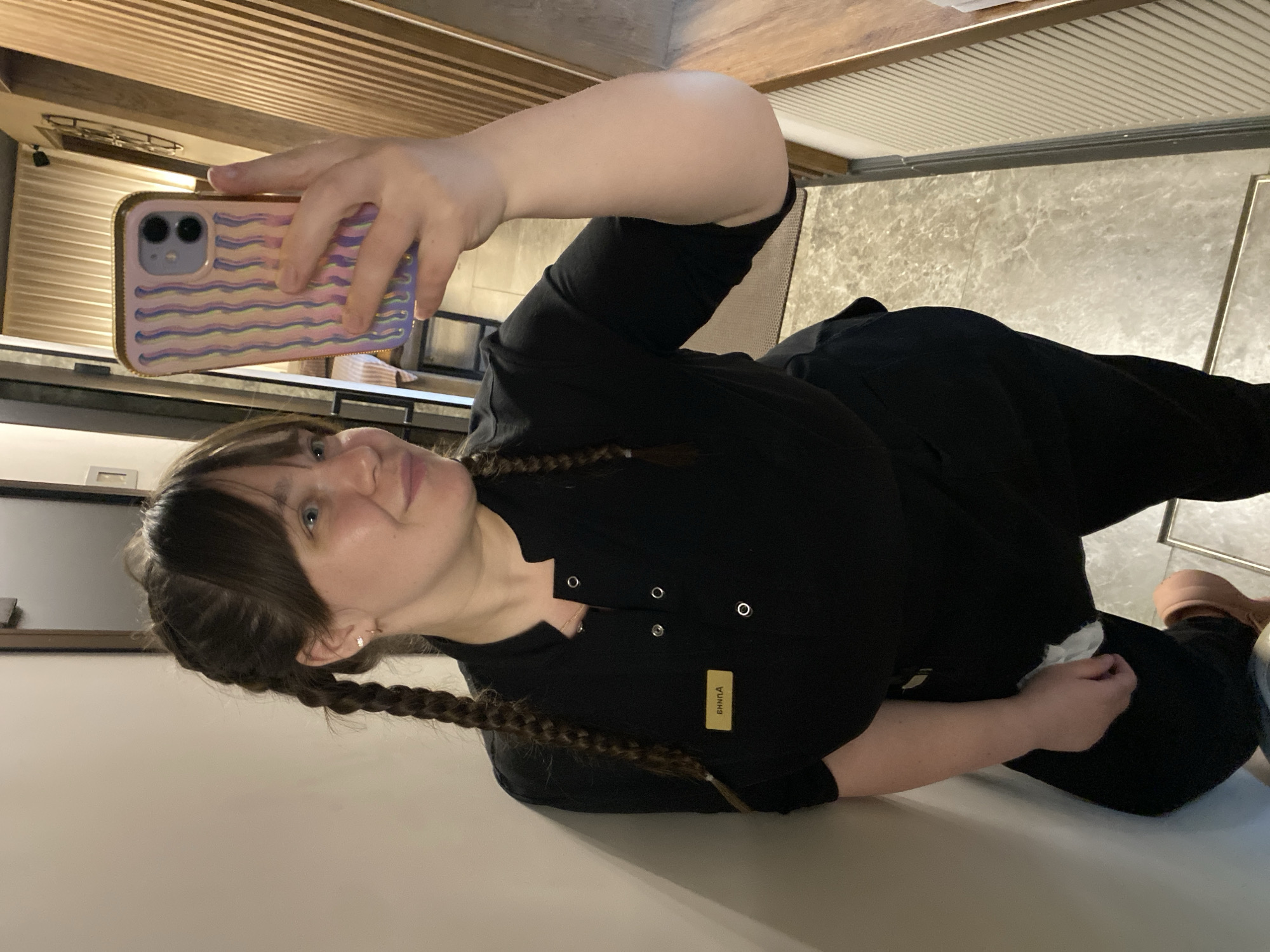 A person with two braids is holding a phone with a striped case, taking a selfie. The person is wearing a black outfit with a label on the front. The background suggests an interior space with paneling and tiled floors. The location cannot be determined as Votkinsk, Russia, from the image alone.
