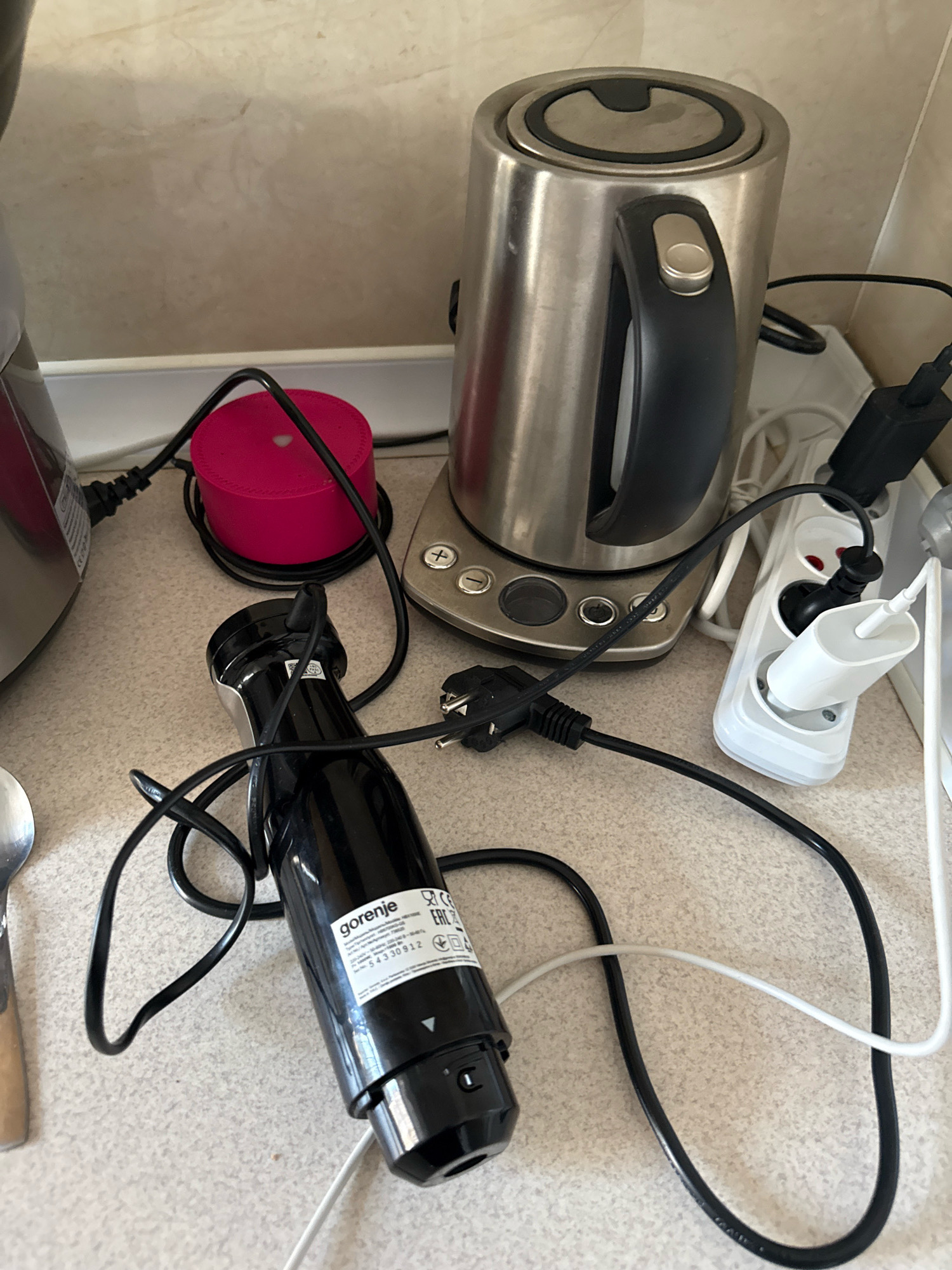 The image depicts a countertop in a kitchen in Chișinău, Moldova. Several kitchen appliances and their power cords are visible. In the foreground, a black immersion blender with the brand name "Gorenje" is present. Its power cord is black and coiled around it. Behind the blender, a stainless steel electric kettle with a black handle and lid is situated. A white multi-socket extension cord is plugged into the wall, with several plugs inserted into it. One plug is a white adapter, and another is a black plug connected to the kettle's cord. A small, round, bright pink device is also visible, with a black power cord plugged into it. The countertop appears to be a speckled beige material, and the background wall has a light beige, possibly tiled or textured finish. The overall scene suggests a domestic kitchen environment with appliances connected to power.