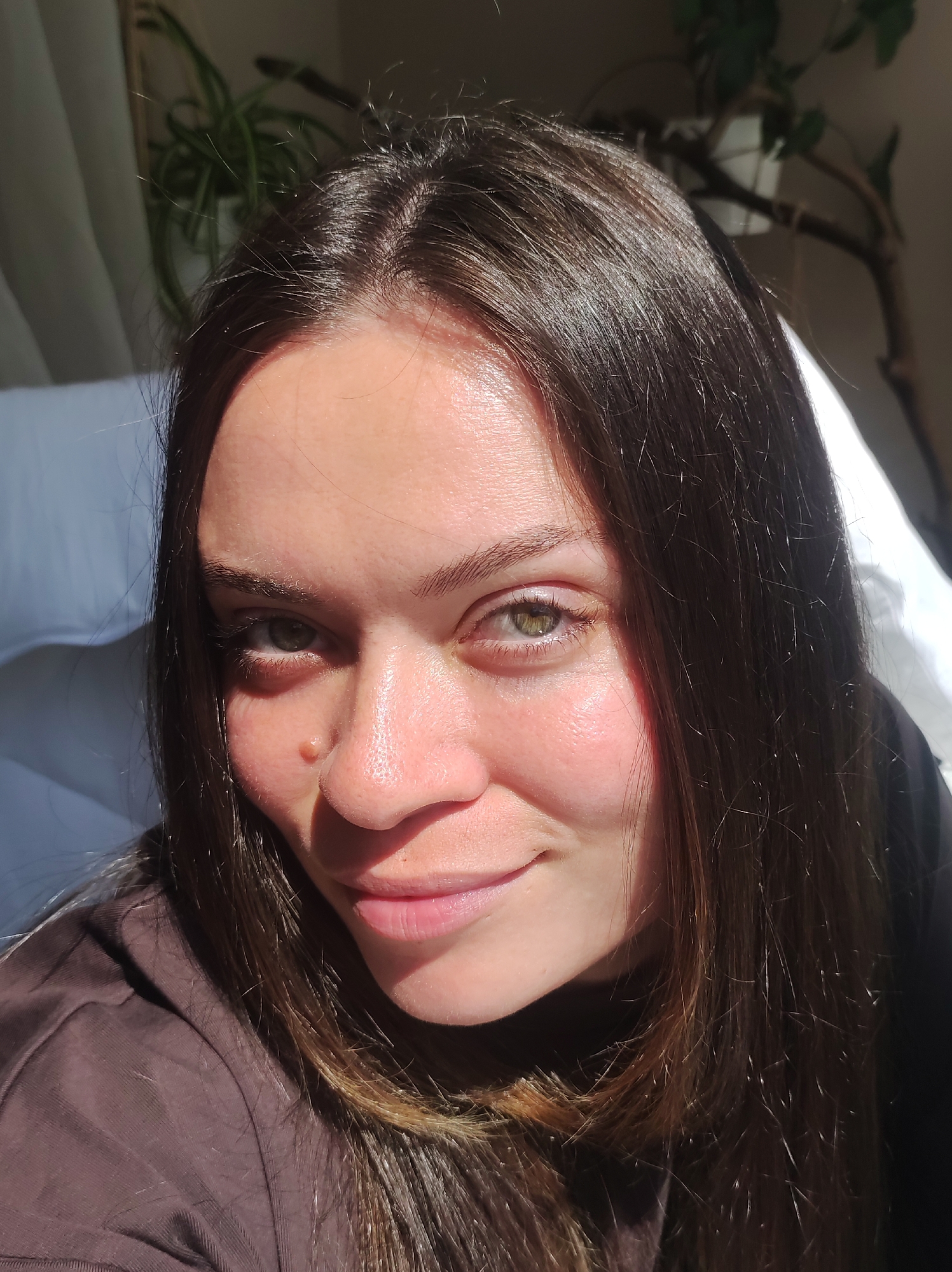 This close-up portrait features a young woman with long, dark brown hair, parted slightly to her left. Her eyes are a striking green or hazel, and she is looking directly at the viewer with a gentle, natural smile. Strong, natural sunlight illuminates her face from the left, creating distinct highlights on her forehead, nose, cheeks, and lips, and emphasizing the natural texture of her skin. A notable mole is visible on the left side of her nose/cheek. The harsh light also casts shadows across the right side of her face and neck.
She is wearing a brown-colored top or shirt. In the background, elements of an indoor setting are visible: blurred green foliage from a houseplant, white fabric (possibly a curtain or bedding), and darker areas, suggesting a room. The strong sunlight indicates it is daytime. The woman appears calm and content. No text is visible in the image. While the location context is Chaikovsky, Russia, no specific visual cues within the image itself confirm this.