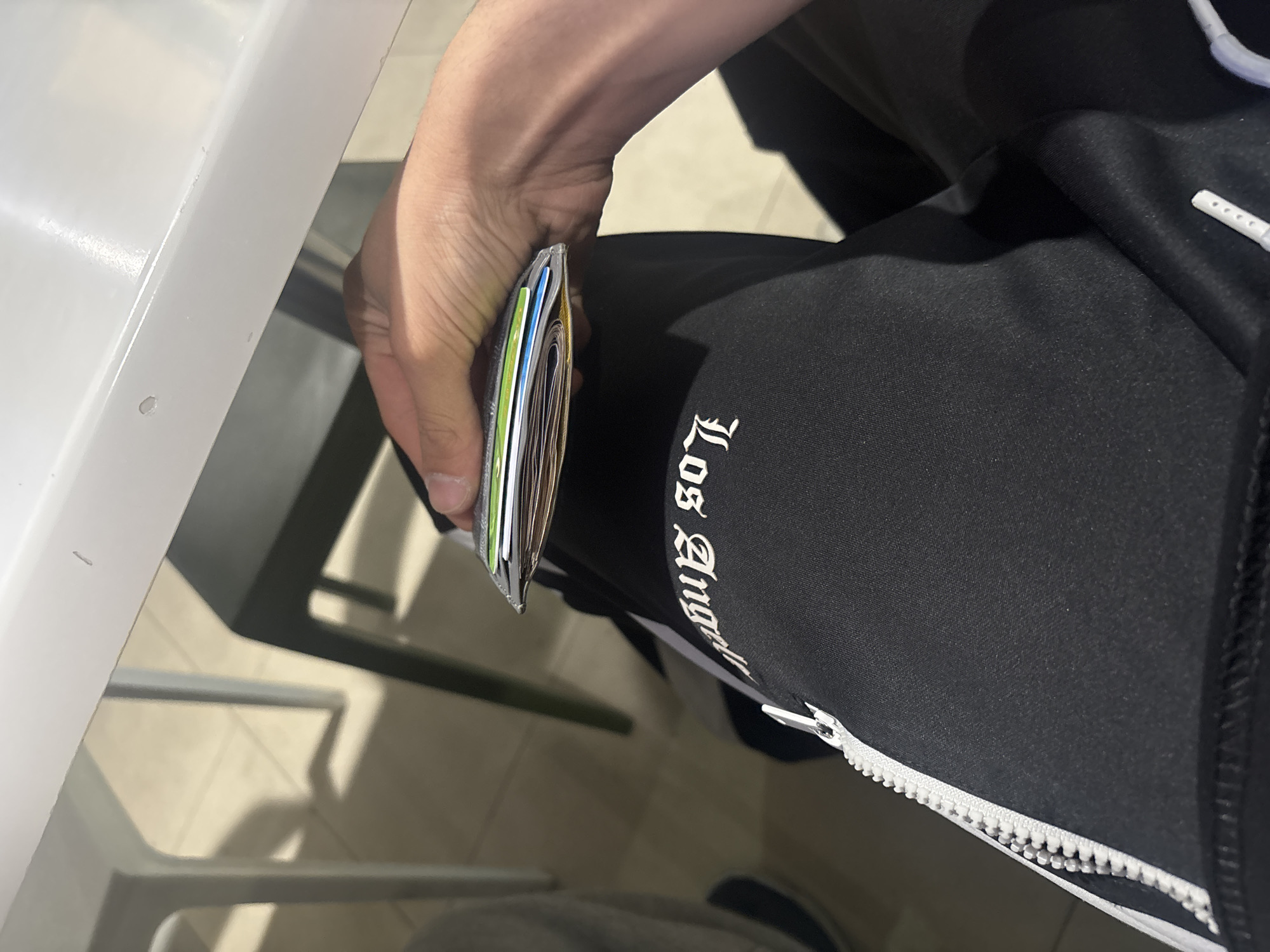 The image presents a close-up, angled view of a person's hand holding a wallet or cardholder in an indoor setting. The primary subject is a right hand with light skin tone, holding a slender, metallic-silver colored wallet. Inside the wallet, several cards with distinct colors such as green, blue, and yellow are visible.
The person is wearing black athletic or lounge pants, likely sweatpants or track pants, which feature a white vertical stripe down the side. Printed on these pants, in a stylized, possibly gothic script, is the partial text "Los Angel," strongly suggesting "Los Angeles" as part of a brand or design. A prominent white zipper is also visible on the side of the pants.
In the background, parts of a white table or counter surface are seen on the left, along with blurred hints of furniture legs and a tiled floor, indicating a casual indoor environment such as a cafe, breakroom, or similar public space.
The activity depicted is the simple act of holding the wallet/cardholder. There are no overt actions to suggest a specific event like a purchase or retrieval of a particular item. The lighting appears to be indoor, but the time of day and weather are not discernible. No emotions can be inferred as no faces are visible. While the provided context suggests Yerevan, Armenia, the image itself does not contain any visual cues to confirm this geographical location.
