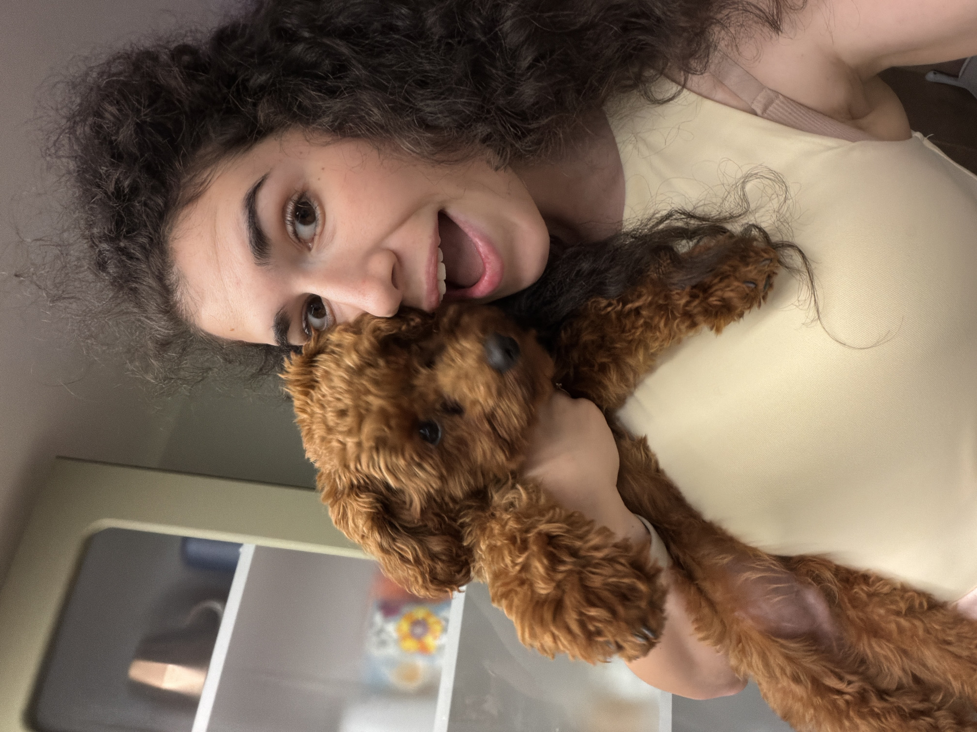 A woman with curly dark hair is holding a small, brown poodle puppy in front of her chest. The woman is looking at the camera with her mouth open, as if she is speaking or laughing. The puppy is nestled against her, with its head peeking over her shoulder. The background appears to be a kitchen or dining area, with shelves containing various items, including what looks like mugs or cups. The lighting is bright, suggesting an indoor setting. The image was taken in Sofia, Bulgaria.