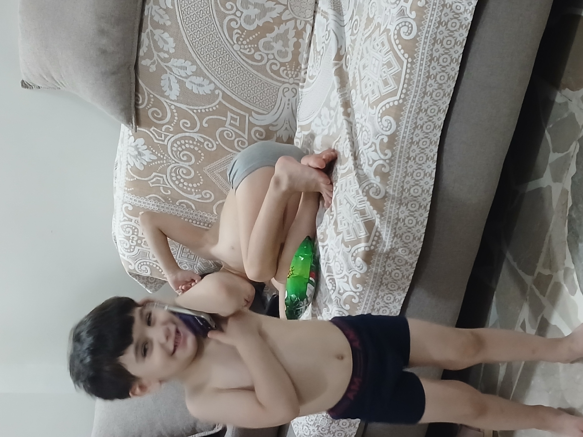 The image shows two young boys on a couch in an indoor setting, likely a living room. The boy in the foreground is on his back, wearing dark blue shorts. He is smiling and appears to be talking on a mobile phone, which he is holding up to his ear. His head is tilted back, and his eyes are wide open. To his left, a light gray cushion is visible.
Behind him, another young boy is lying on his stomach, curled up with his knees drawn to his chest. He is wearing light gray underwear. A green, shiny object, possibly a toy or a snack wrapper, is positioned near his feet. The couch is covered with a patterned beige and white fabric.
The lighting suggests it is daytime. The overall mood of the image appears to be relaxed and playful, with the boy in the foreground expressing happiness. The scene is domestic, with the couch and patterned fabric indicating a home environment. There is no visible text in the image.