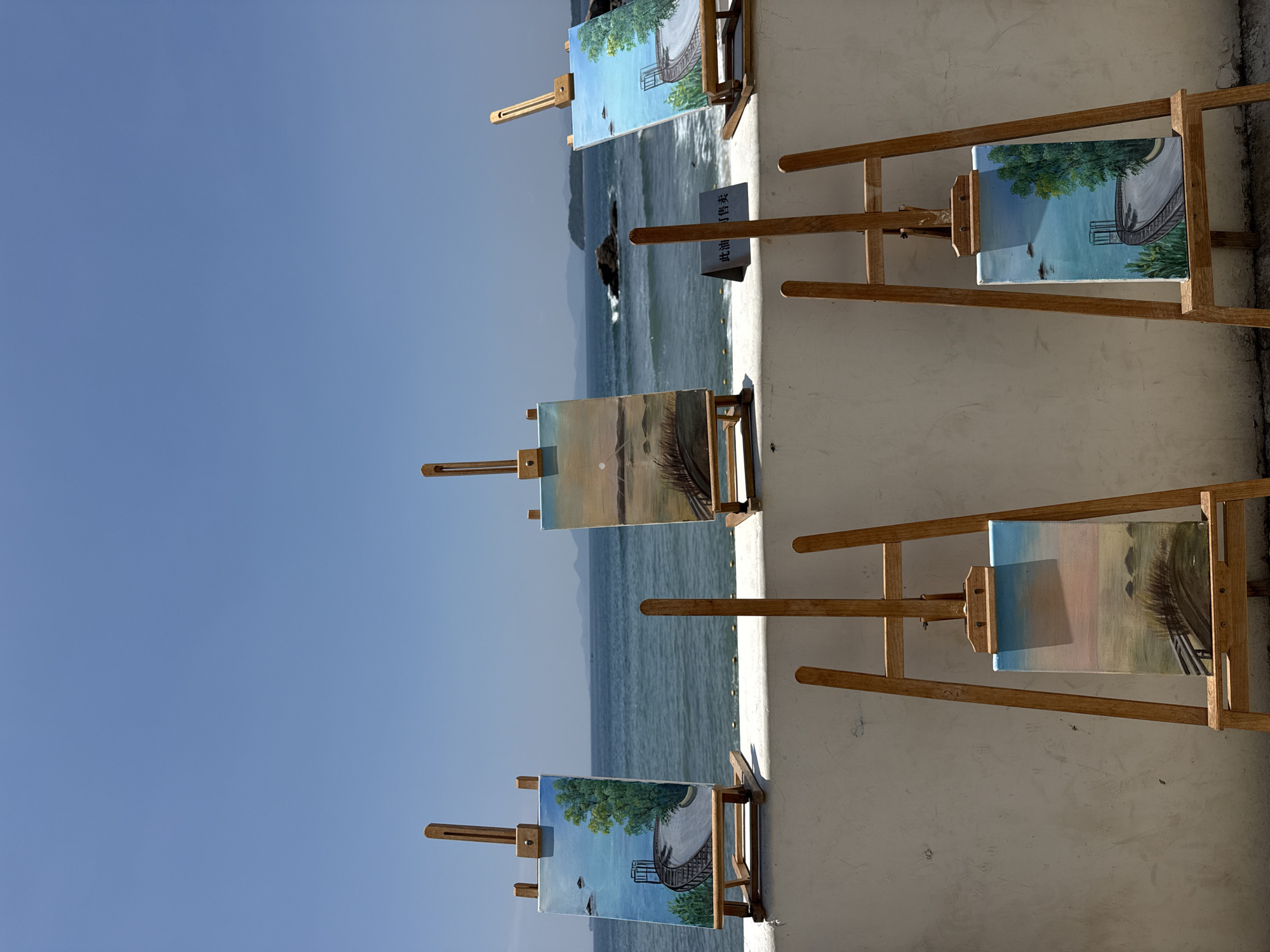 In the city of Longgang District, China, a scenic outdoor art display features several small paintings placed on wooden easels. The paintings, all depicting coastal or seaside landscapes, are arranged along a white, textured wall. The ocean is visible in the background, with gentle waves and a clear blue sky overhead. Islands and distant mountains are present on the horizon. The paintings themselves showcase various scenes, including a pathway lined with trees, a sunset over the water, and a view of the sea. One painting has a small sign in front of it with Chinese characters. The overall impression is one of a tranquil, artistic setting by the sea.