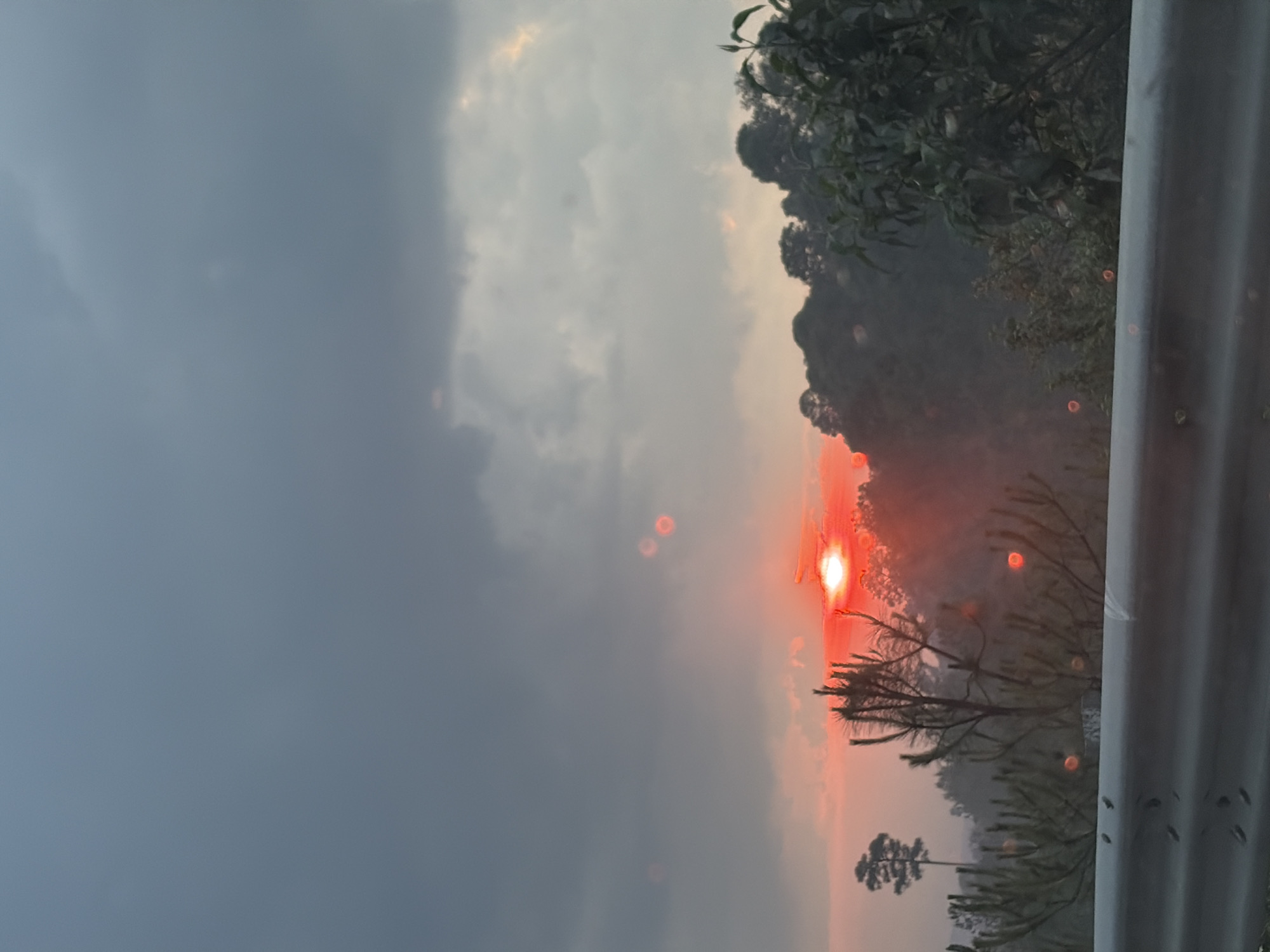 The image shows a landscape at sunset in a location possibly near Chalco de Díaz Covarrubias, Mexico. The sky is divided between heavy, dark grey clouds on the left and a lighter area with streaks of orange and yellow where the sun is setting on the right. A bright red sun is partially obscured by the horizon line and silhouetted trees. Some vegetation is visible along the horizon line. In the foreground, there is a grey metal guardrail. There are some lens flares visible in the image.
