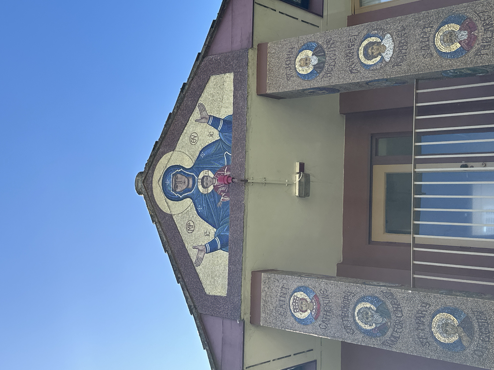 An exterior, low-angle, forensic view captures the upper portion of the Dormition of the Mother of God and St Cedd Orthodox Church, located in Maldon, United Kingdom, under a clear, bright blue sky.
The central feature is a large, triangular mosaic adorning the gabled facade. This mosaic depicts the Virgin Mary, known as the Mother of God, in a blue robe with a golden halo, hands raised and palms open. In front of her is the infant Jesus Christ, dressed in red garments with a golden halo. Greek abbreviations "MP OY" (Μήτηρ Θεού, Mother of God) are visible near the Virgin Mary, and "IC XC" (Ἰησοῦς Χριστός, Jesus Christ) are visible near the infant. The mosaic is composed of numerous small, uniformly shaped tiles, primarily gold for the background, blue, and various skin tones.
Below the main mosaic, two rectangular pillars support an overhang. Both pillars are also covered in mosaics, featuring circular portraits of saints and religious figures.
The left pillar, from top to bottom, displays:
1. A crowned female figure identified by text as "SAINT CATHERINE".
2. A bearded male figure identified by text as "ST PROCOP FOOL FOR CHRIST".
3. A female figure identified by text as "ST MARY OF EGYPT".
The right pillar, from top to bottom, displays:
1. A bearded male figure identified by text as "ST JOHN THE DIVINE".
2. A winged figure identified by text as "ARCHANGEL RAPHAEL".
3. A bearded male figure identified by text as "SAINT PHILIP".
All figures on the pillars possess golden halos. The building's walls feature reddish-brown and cream/beige sections. A white-railed balcony is visible in the lower frame, in front of a light-colored door and a dark-paned window. A white light fixture is mounted centrally on the underside of the overhang. A small, red pipe and white electrical conduit are affixed to the wall near the left side of the main mosaic. The scene is brightly illuminated by direct sunlight.