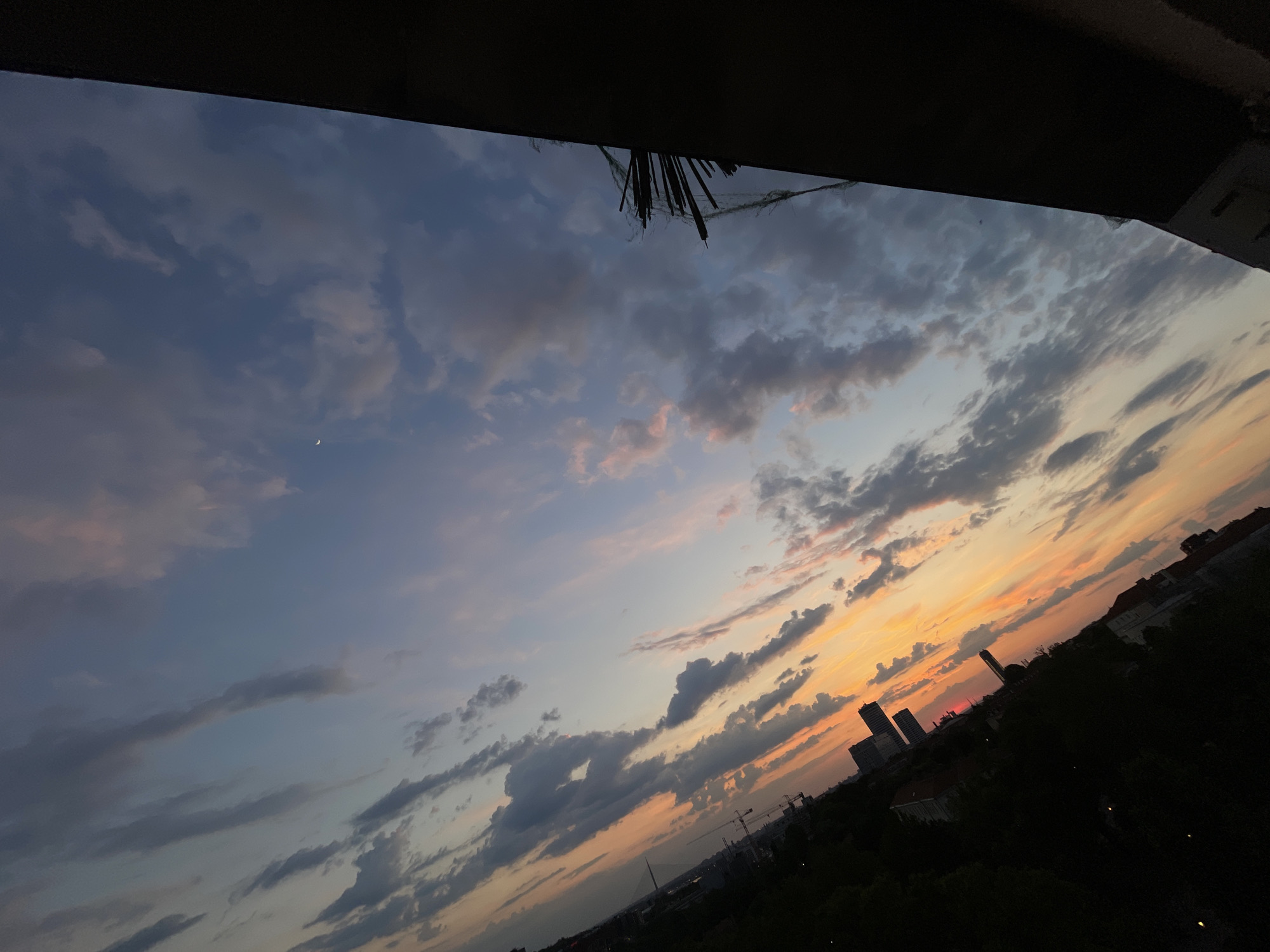 View from an elevated position in Vracar Urban Municipality, Serbia, at sunset. The sky is a mix of blue and orange, with clouds scattered across it. A sliver of the moon is visible. Below, there are silhouettes of buildings and trees. Construction cranes are visible in the distance. The foreground is dark, obscuring details. A structural overhang is present at the top of the frame.