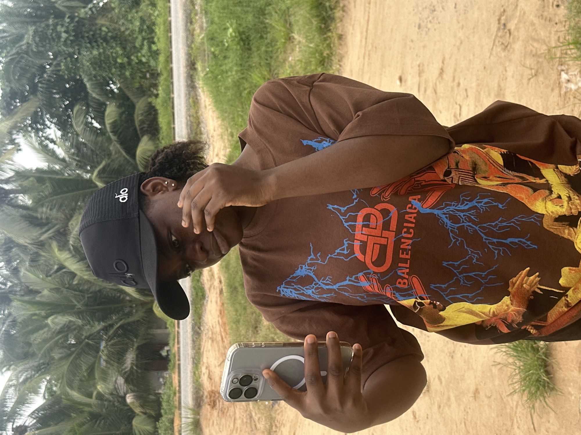 A young Black woman, wearing a black trucker hat and a brown t-shirt with a graphic print, stands outdoors and holds a smartphone in her left hand, taking a selfie. The woman has her right hand raised to her face, touching her eye. She is looking at the phone screen, which is not visible. The t-shirt features a large graphic with blue lightning bolts and text that reads "BALENCIAGA". The woman is positioned in the foreground against a backdrop of palm trees and greenery. The ground appears to be sandy and dirt. The location is identified as Ganyingbo, Nigeria.