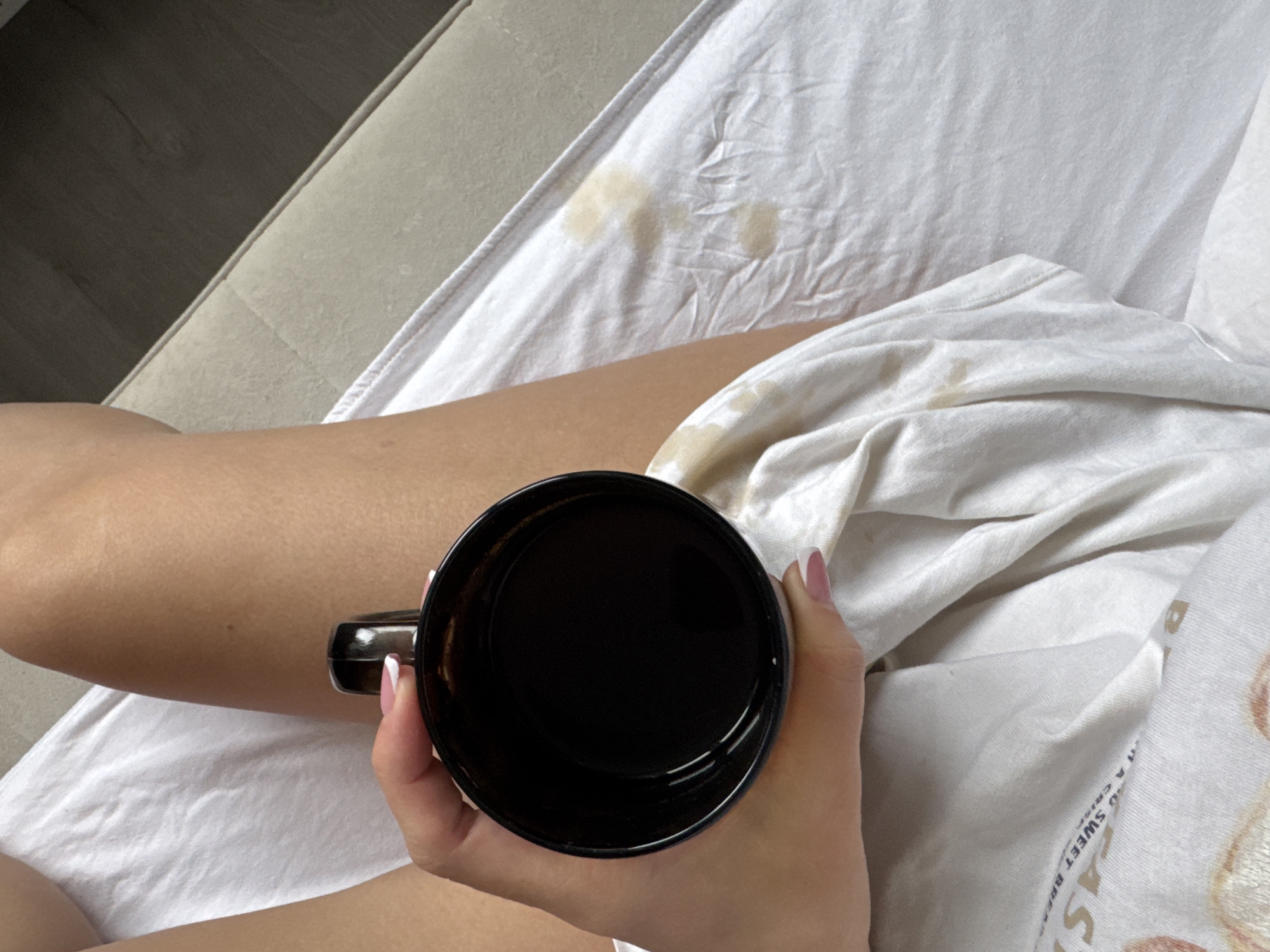 An indoor, overhead shot captures a person's leg and hand holding a black mug, seemingly filled with a dark liquid. The person's bare leg occupies the left and central-left portion of the frame, resting on or against light-colored fabric, possibly a sofa or bed. Their hand, with nails painted in a French manicure style, grips the black mug from below, with the mug's opening facing upwards and appearing almost full.
The dominant visual element is a large expanse of white or off-white fabric, which is noticeably stained with multiple light brown, irregular marks, consistent with spilled coffee or tea. These stains are concentrated towards the upper center and lower right of the image. The fabric itself is rumpled in places, suggesting it could be bedding or a soft furnishing. In the background to the far left, a dark wooden surface, likely a floor, meets a lighter, possibly carpeted or wall area, at an angle.
The activity depicted is the act of holding a beverage, possibly just after or during a spill, given the prominent stains on the fabric. The scene conveys a casual, relaxed atmosphere, perhaps in a domestic setting like a bedroom or living room. Visible text on the white fabric in the lower right includes partially legible words such as "A SWEET BREA" and "...AST", suggesting "BREAKFAST", and "A CRISP...". No specific time of day or weather cues are evident. The setting is identified as Chaikovsky, Russia, based on provided context, though no explicit visual cues within the image confirm this location.