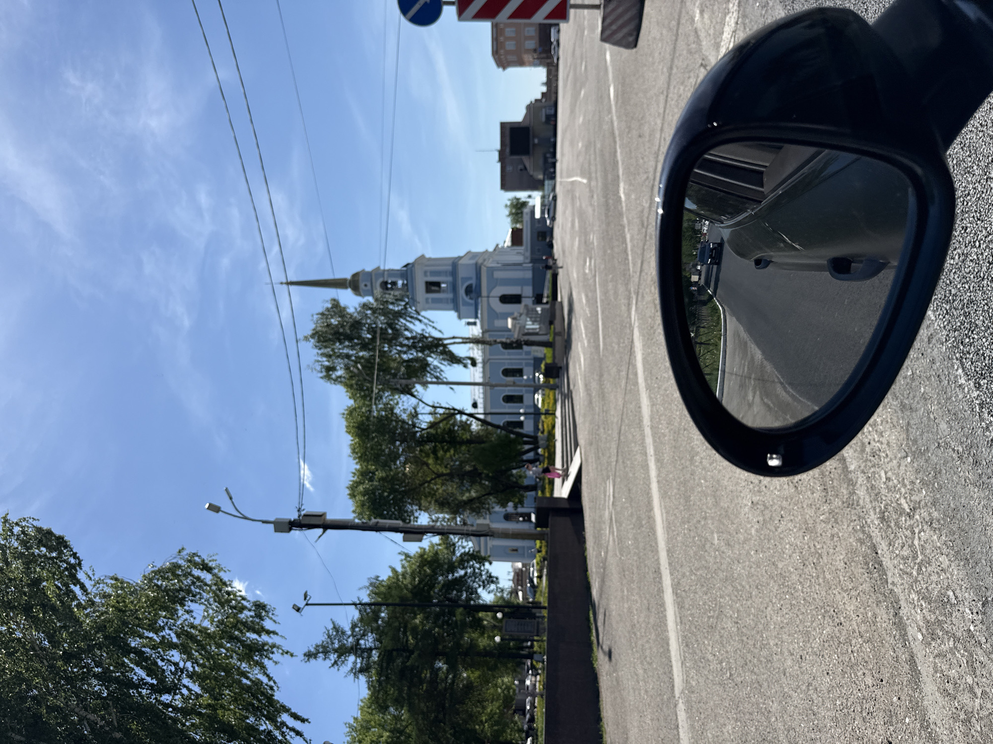 The image shows a view from a car on a city street, oriented vertically. The car's side mirror is visible in the right foreground, reflecting a road behind the car with other vehicles. The primary subject is the Annunciation Cathedral in Votkinsk, Russia, visible in the middle ground. A crosswalk and trees line the street leading to the cathedral. Streetlights and overhead wires are present. Other buildings are further back in the background. The sky is blue with wispy clouds. The road surface is asphalt, showing wear.
