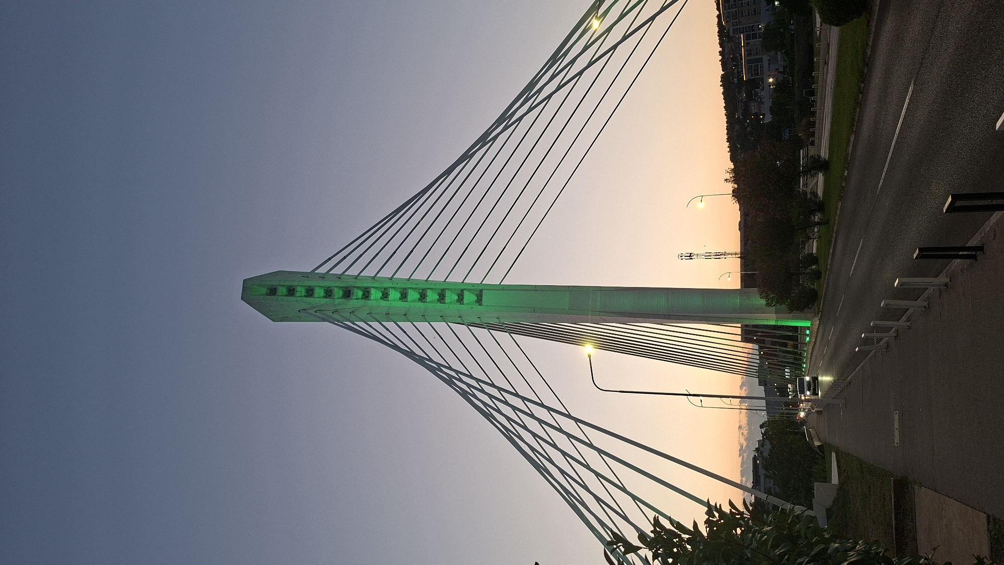 The image shows the Millennium Bridge in Podgorica, Montenegro, at dusk. The bridge's central pylon is illuminated with green lights. The sky transitions from a deep blue at the top to a pale orange near the horizon, indicating the setting sun. A road runs alongside the bridge, with cars visible in the distance. A sidewalk is to the left of the road, with trees and foliage bordering it. The scene is urban, with buildings visible on the hills in the background.
