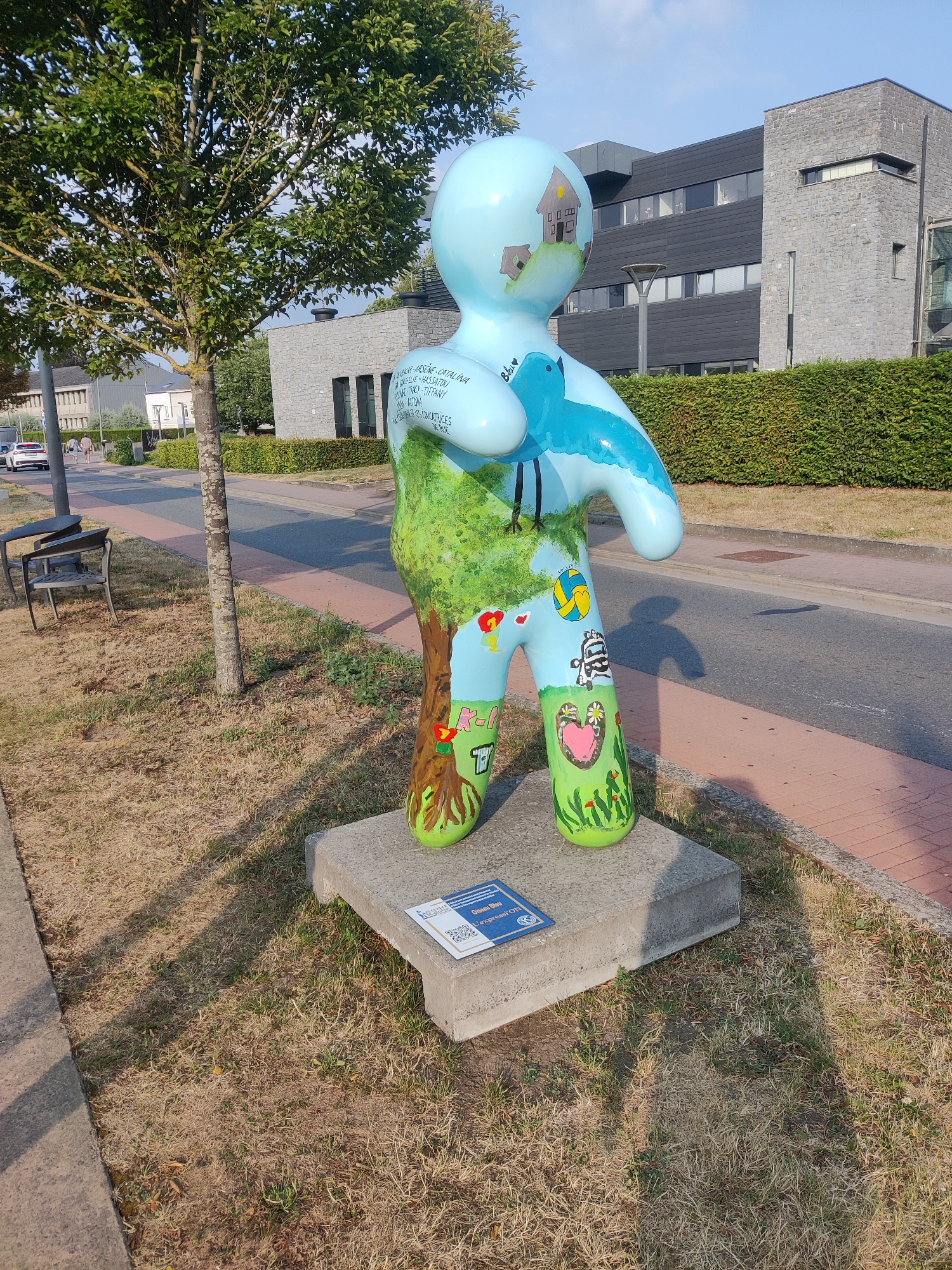 A light blue, anthropomorphic sculpture painted with various images is displayed on a concrete base in front of a building with dark grey siding and a stone facade in Marche-en-Famenne, Belgium. The sculpture is painted with a tree on its torso, a bird on its chest, a house on its head, and various designs on its legs, including a heart and a zebra. The sculpture is surrounded by dry, brown grass, with a paved walkway and a street visible in the background. A thin tree with green leaves stands to the left of the sculpture, and a portion of a building with multiple windows is visible behind it. The sky is clear and blue. There are no people in the immediate vicinity of the sculpture.