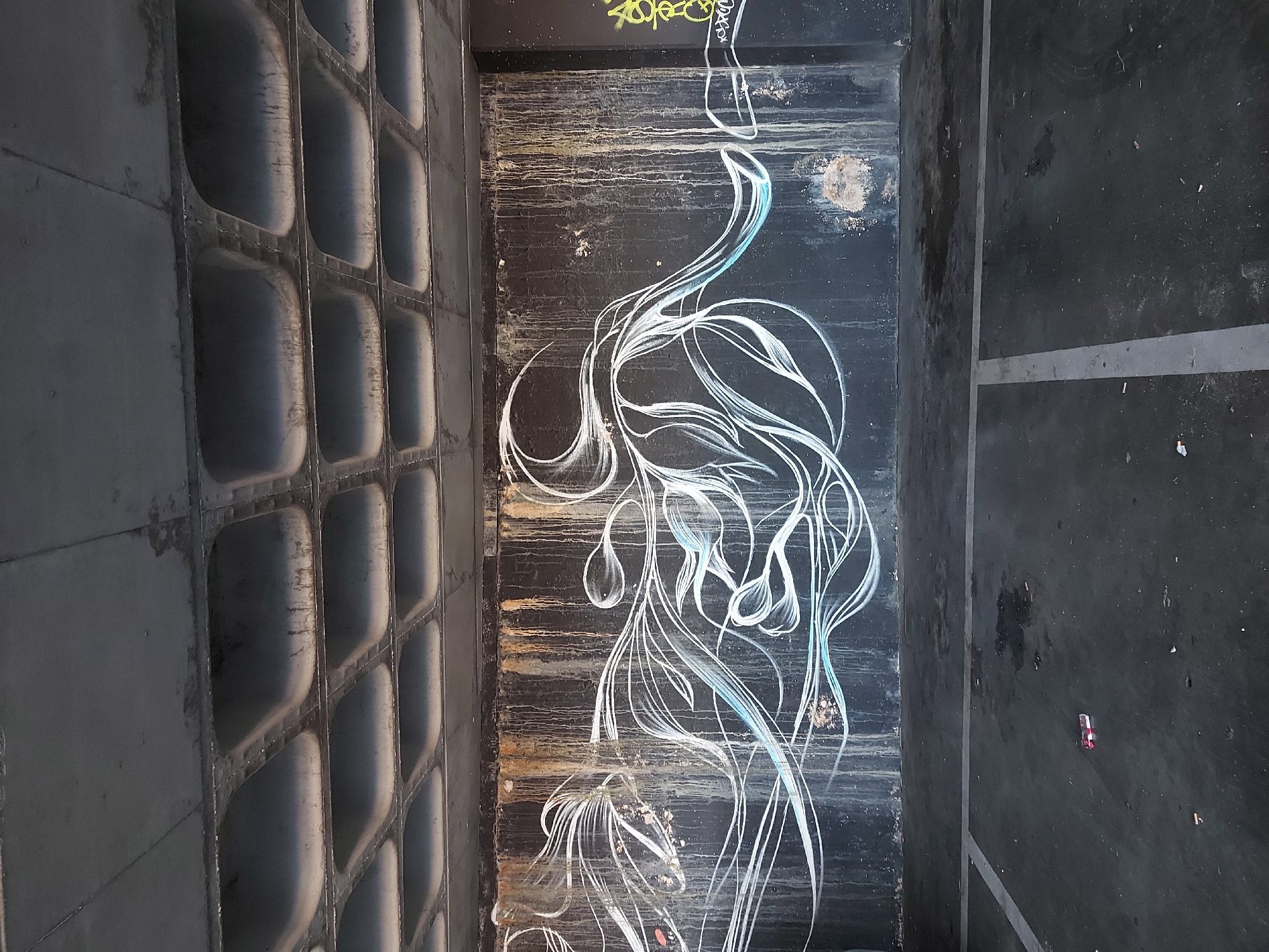 The image depicts an urban scene in Figueira da Foz, Portugal. A section of a concrete wall with a grid-like pattern of hollow, rounded rectangular cells is visible on the left. Adjacent to the wall is a dark, possibly painted, surface featuring white graffiti art resembling stylized floral or organic forms. The graffiti includes hints of blue shading. The surface shows signs of wear and rust. To the right of the graffiti surface is a dark, paved area with a few small objects scattered on it, including what appears to be a small, rectangular object (possibly a cigarette butt or small wrapper). A thin, white line runs across the paved area. The photograph is taken from an elevated or angled perspective.