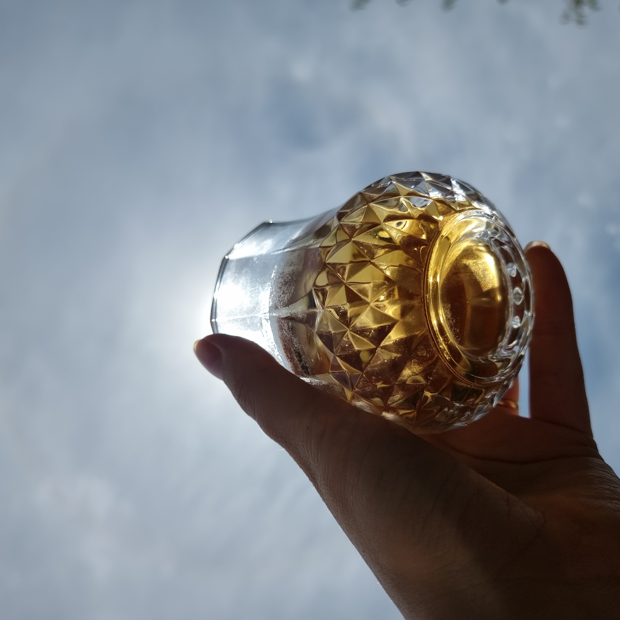A hand holds up a clear glass tumbler filled with a golden liquid, possibly beer or whiskey, against a hazy sky with subtle cloud formations. The glass has a faceted, diamond-like pattern that reflects the sunlight filtering through the sky, creating bright highlights. The sun is partially obscured by the rim of the glass, casting a lens flare. The background is primarily the sky, with some faint greenery possibly from trees visible at the edges, indicating an outdoor setting. The image appears to be taken in Dunnae-myeon, South Korea, given the provided context.