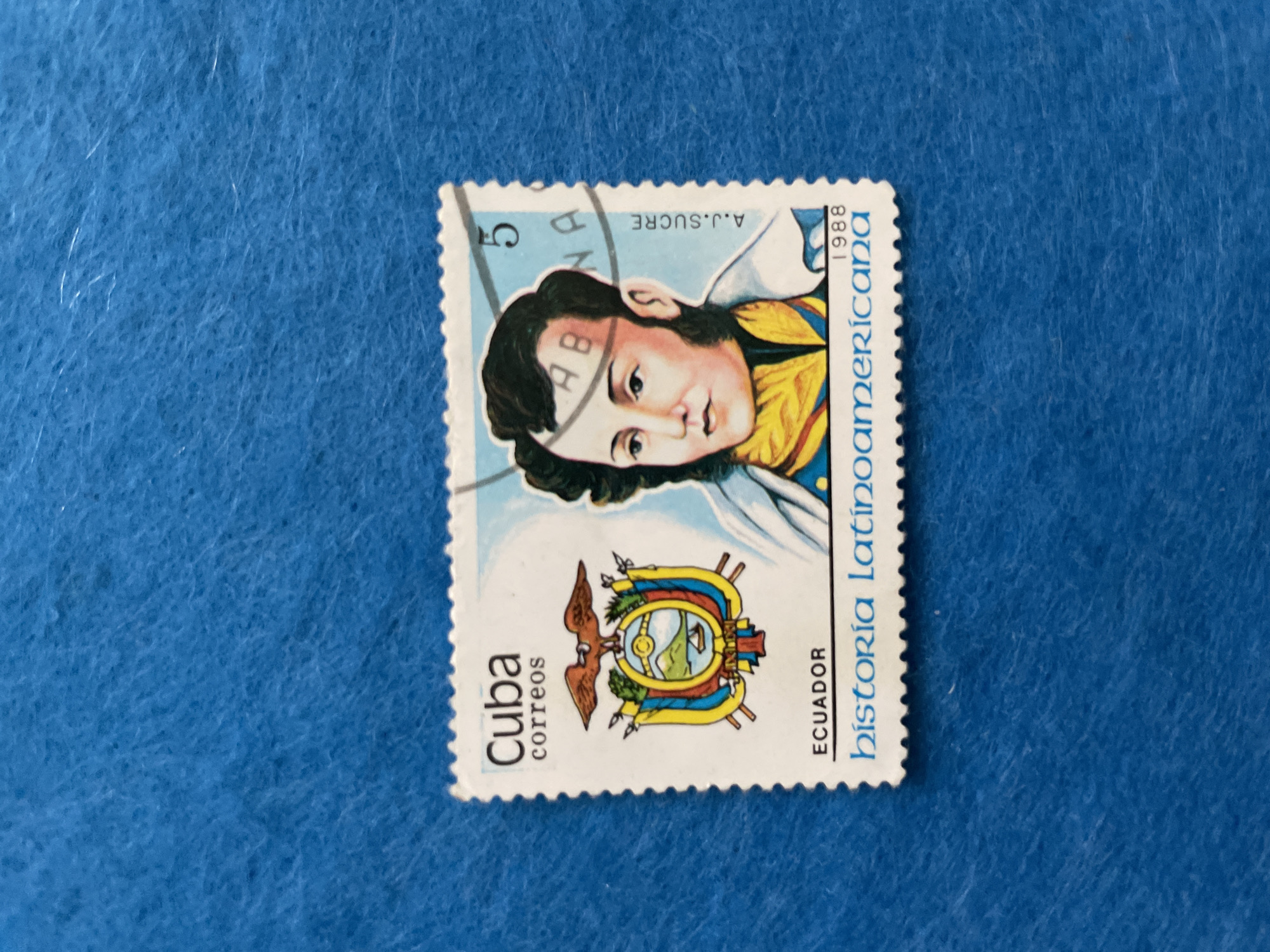 A postage stamp featuring a portrait of Antonio José de Sucre, a Venezuelan independence leader, is placed on a blue textured surface. The stamp is white with colored illustrations. The text "Cuba correos" is printed at the top left, with the Ecuadorian coat of arms below it. To the right of the coat of arms is Sucre's portrait, with the number "5" in a diagonal line across his forehead. Text in a script font reads "A.J.SUCRE" vertically on the right side of the stamp. Below the portrait, "ECUADOR" is printed, followed by "historia latinoamericana" and "1988". The blue surface appears to be fabric, possibly felt. The location in the city of Perm, Russia, is not ascertainable from the image.