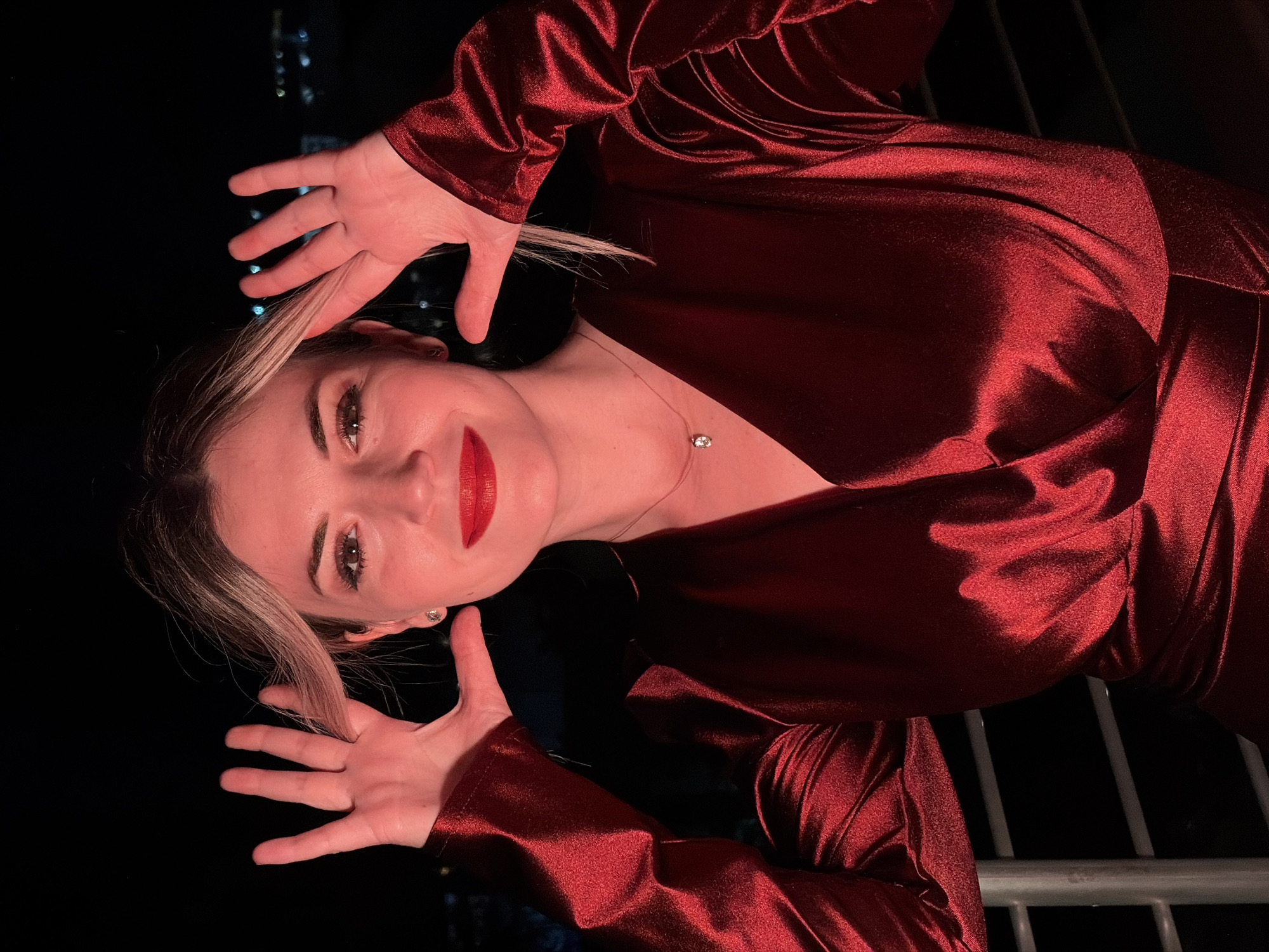 A woman with blonde hair and red lipstick poses with her hands framing her face in front of a dark background, possibly a city at night. She is wearing a shimmering red dress. A railing is visible in the lower left corner of the image. The scene appears to be outdoors, possibly on a balcony in Budva, Montenegro, given the faint lights of the city in the distance.