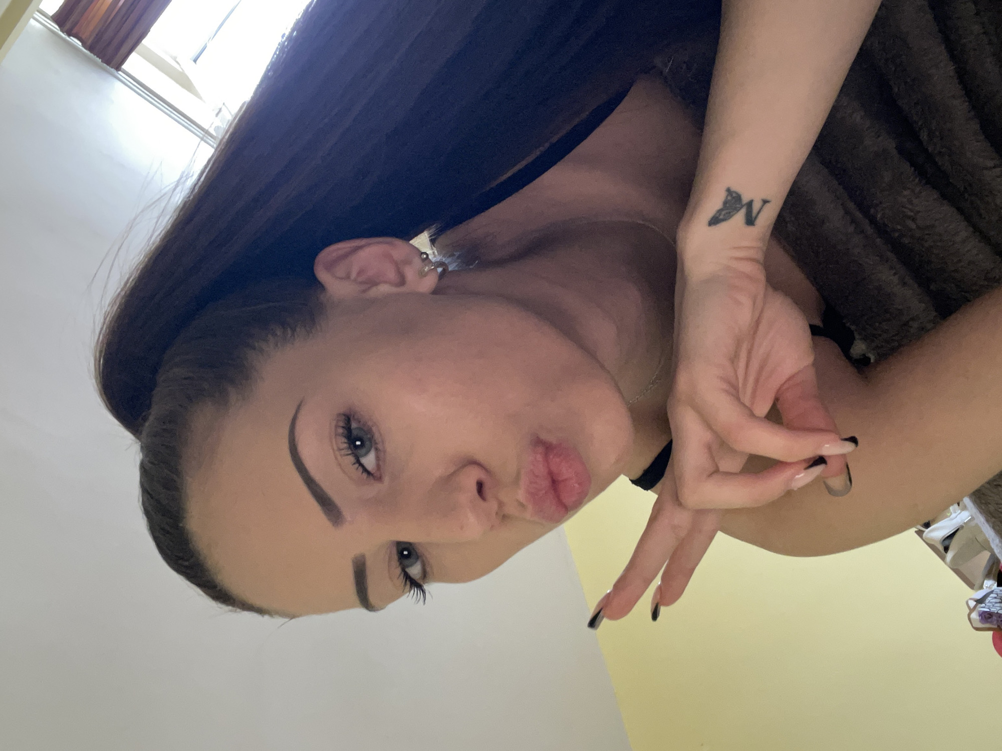 A young woman poses for a selfie indoors. She has long dark brown hair and light eyes. Her lips are pursed slightly. There's a butterfly and the letter "N" tattoo on her left wrist. She is wearing a choker necklace. The location appears to be a residential room, with yellow walls and natural light coming from a window in the upper left corner. The location in the city is unknown.