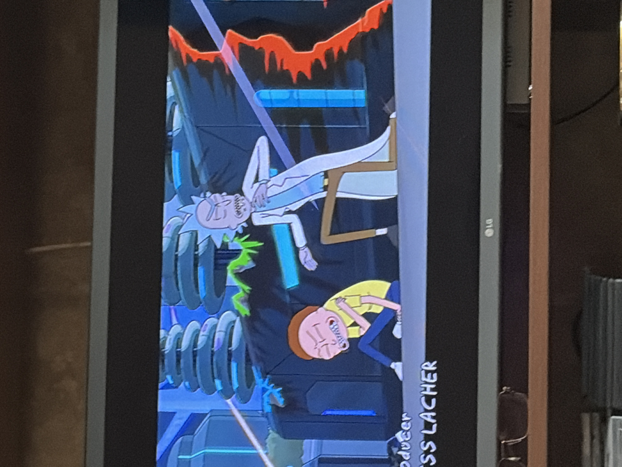In a scene from the animated show Rick and Morty, Rick Sanchez, a tall, bald man with green skin and messy hair, stands in a laboratory setting. He is wearing a lab coat and appears to be in distress, clutching his chest. Morty Smith, a young boy with spiky brown hair, sits on the floor with his eyes closed and his arms wrapped around himself, also seemingly in pain. The background features futuristic machinery and a swirling vortex of energy. The bottom of the screen displays credits, including "ODUCER" and "SS LACHER." The scene is viewed on a television screen, with the bottom edge of the television displaying an LG logo.