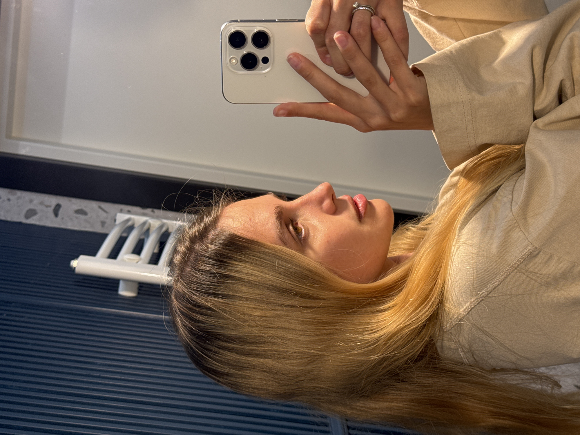 A person is holding a white iPhone in their hands in front of a mirror, taking a selfie. The individual is wearing a beige top and a ring on their finger. Their blonde hair is visible along with a portion of their face. The background includes a tiled surface and a white towel rack. The location is Unknown, Unknown.
