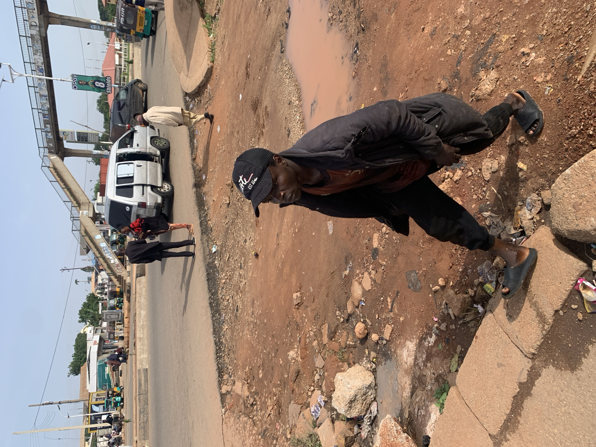 The image captures a street scene in Gura Top, Nigeria, likely a commercial or transportation hub, indicated by the presence of various vehicles and a pedestrian overpass. In the foreground, a man wearing a black jacket, a red t-shirt, black pants, and black sandals walks on a dirt path strewn with rocks and debris. He is wearing a black baseball cap. To his left, a section of paved curb is visible, also with some litter. In the mid-ground, a silver minivan with its doors open is parked on a paved road. Behind it, a black SUV and several auto-rickshaws, commonly known as Keke Napep, are present. Further in the background, a pedestrian bridge spans across the road, with advertisements displayed on its pillars. Other people are seen near shops and on the road. The environment appears somewhat dusty and unpaved in areas, typical of many urban environments in Nigeria. The lighting suggests it is daytime.
