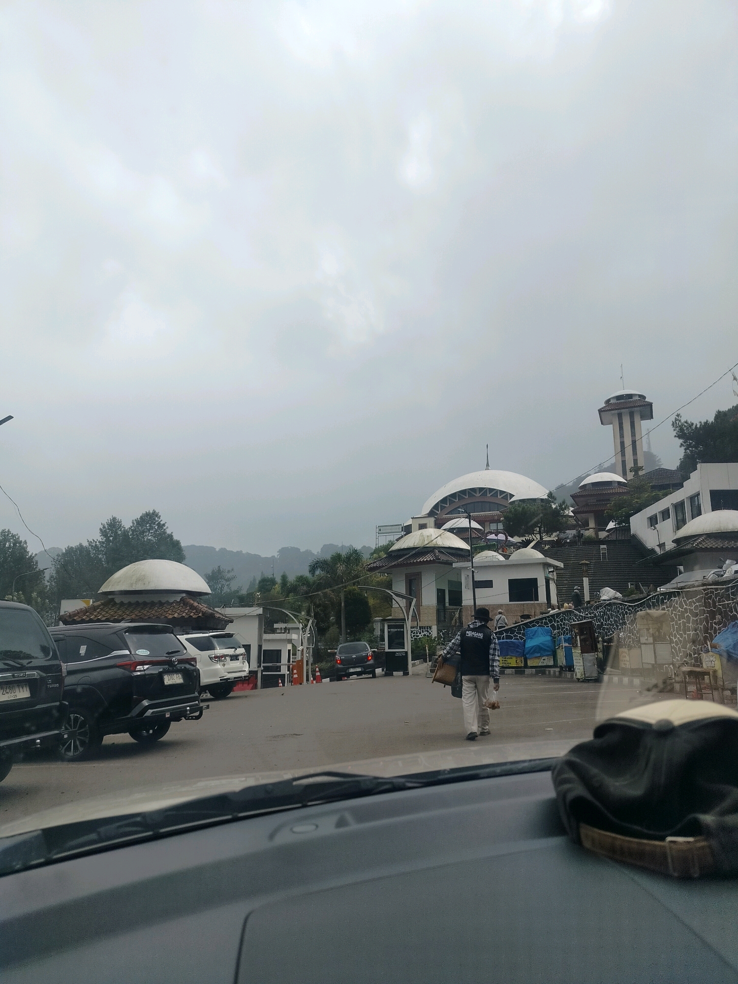 The image captures a frontal view from within a vehicle, looking out at a street scene in Cisarua, Indonesia, under an overcast sky. In the foreground, the dashboard and a dark baseball cap are visible. The middle ground shows a line of parked vehicles, including a black SUV and a white SUV. A person wearing a black vest with "PEDAGANG" (Indonesian for "merchant") written on the back is walking away from the camera, carrying bags. Behind the parked cars and the pedestrian, several buildings are visible, including structures with domed roofs and a tall tower. The architecture suggests a public or tourist area, possibly a religious site or a recreational complex. The background features lush green hills and trees, characteristic of the mountainous region of Cisarua. The overall atmosphere is subdued due to the cloudy weather.