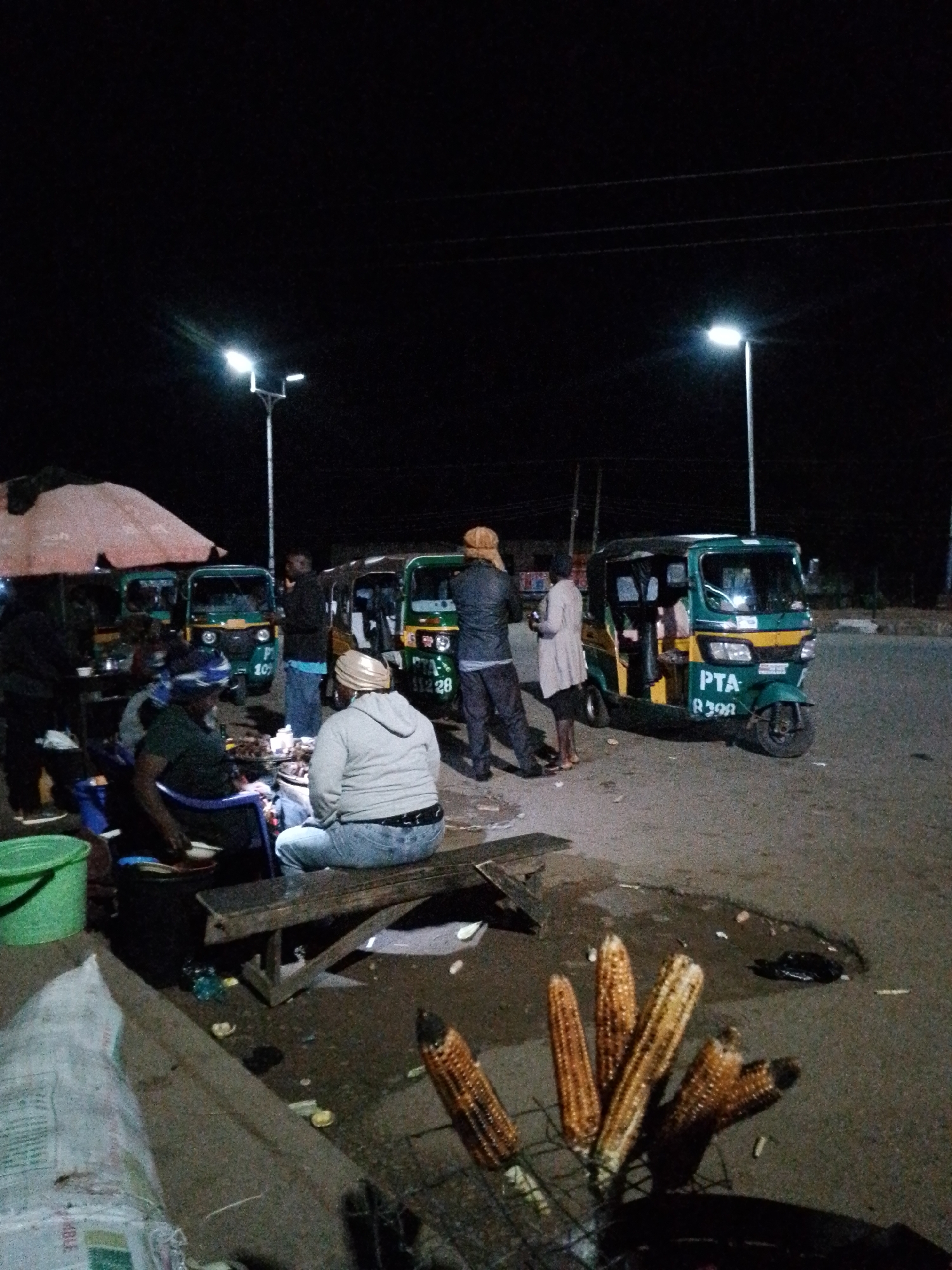 This is a night-time scene in Bukuru, Nigeria, likely a roadside vending area. Several auto-rickshaws, painted green and yellow with "PTA" markings, are parked on a paved surface. People are gathered around food stalls, with some sitting on benches and others standing. Grilled corn on the cob is visible on a rack in the foreground, suggesting food is being prepared and sold. Streetlights illuminate the scene, casting long shadows. The overall atmosphere appears to be one of an active, albeit dimly lit, commercial hub.