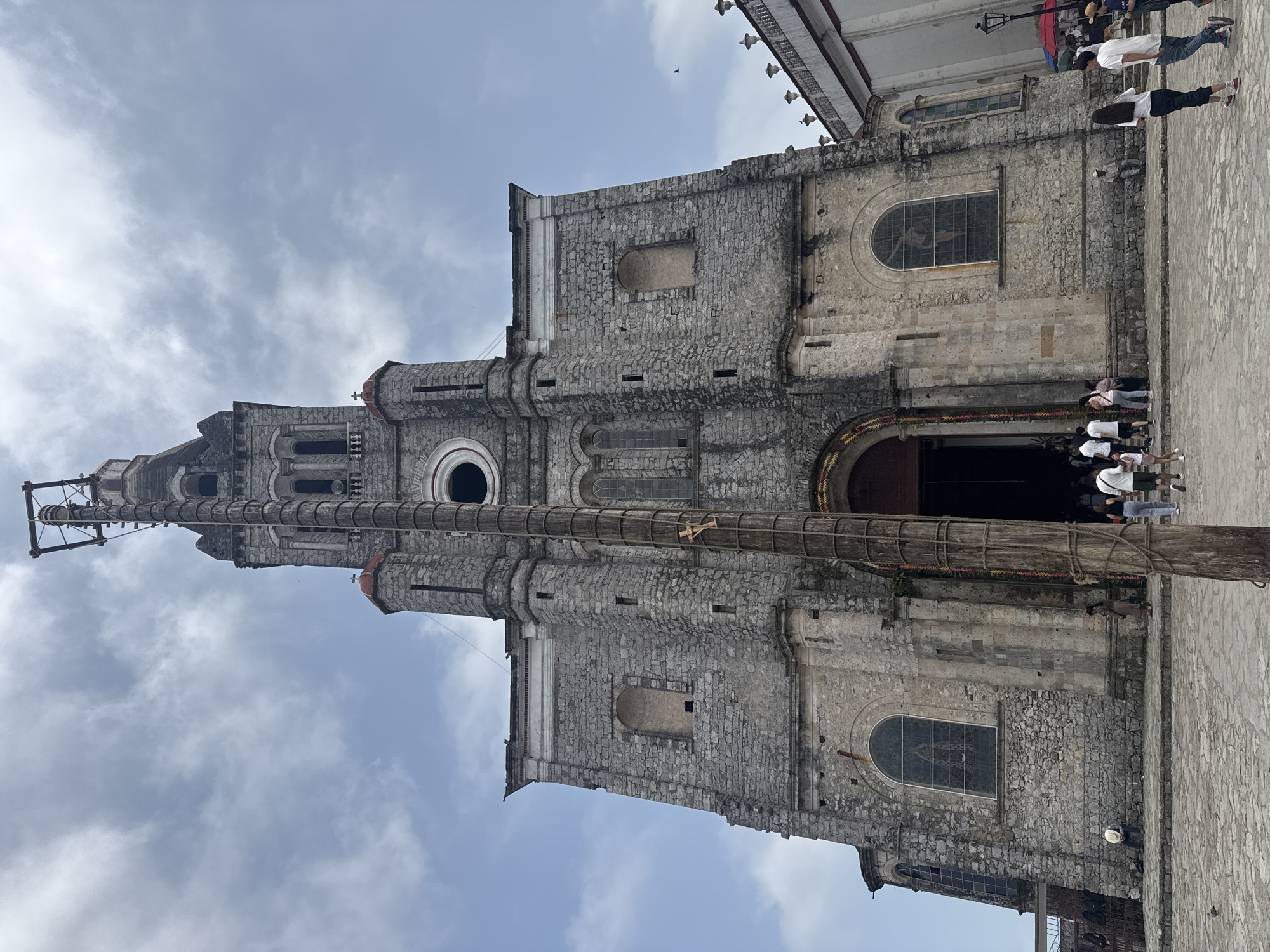The image displays a grand, historic stone church, likely in Cuetzalan, Mexico, under a partially cloudy sky. The church is constructed from grey, rough-hewn stone blocks, featuring a detailed facade with multiple arched windows and a prominent, tall bell tower on its left side. Intricate architectural details, including round windows and decorative carvings, adorn the structure.
In front of the church, a very tall and thick wooden pole stands upright, stretching high above the church's entrance and much of its facade. This pole is characteristic of the "Danza de los Voladores" (Dance of the Flyers) ritual, a traditional Mesoamerican performance. It has visible rope-like bindings and a wooden framework near its top, suggesting preparation for or part of this cultural ceremony.
Several people are gathered in the paved, cobblestone plaza in front of the church and around the base of the wooden pole. They appear to be spectators or visitors, dressed in casual clothing, observing the scene. The church's main entrance, a large dark archway, is visible behind the pole, hinting at an ongoing or impending event.
The time of day is daytime, with soft, diffused light indicating an overcast or partly cloudy sky. The overall atmosphere suggests a public gathering at a significant historical and cultural site. No discernible text is visible on any part of the church or its surroundings.