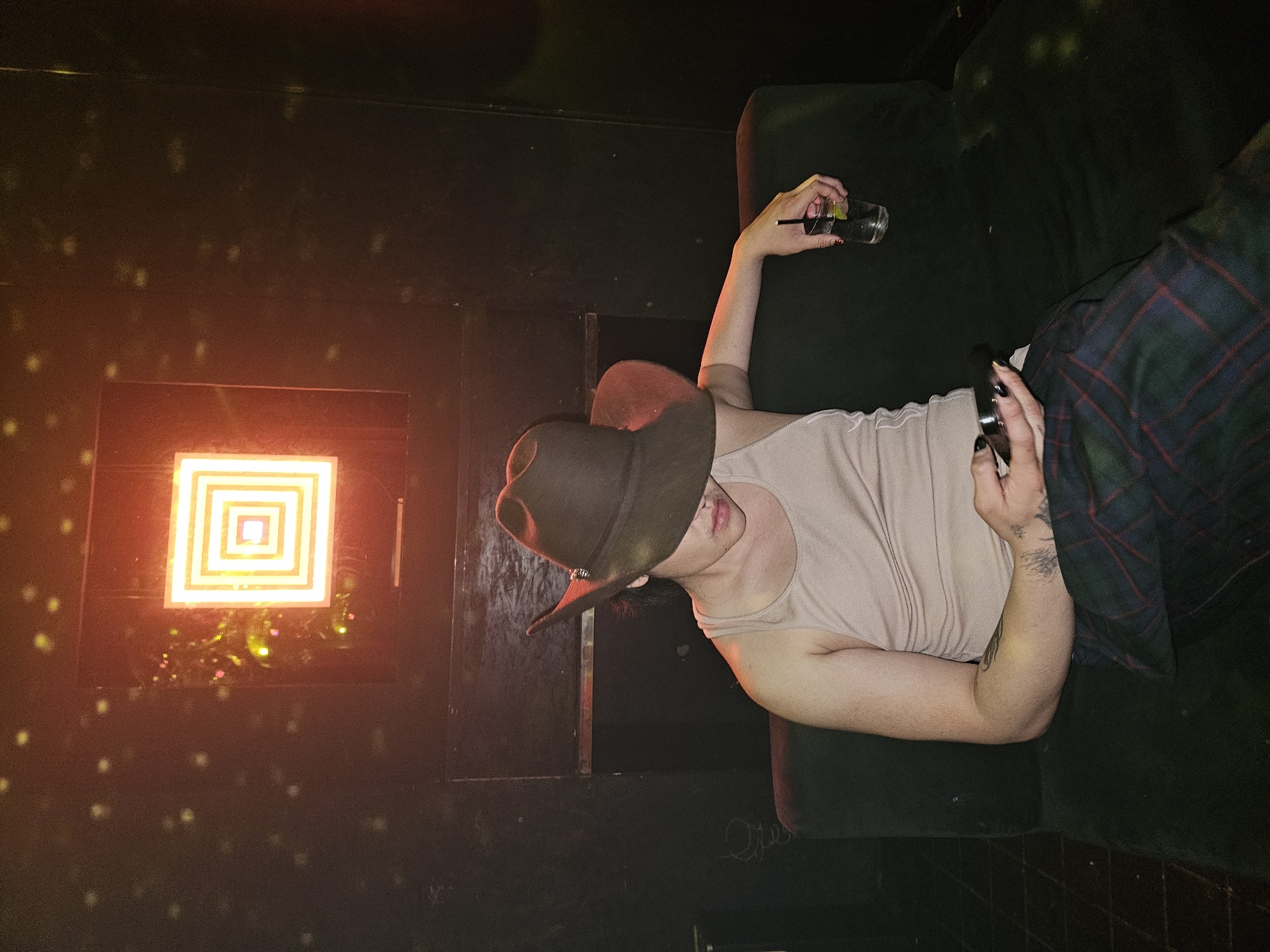 A person lies on a dark green velvet-like couch. They wear a brown cowboy hat that obscures most of their face, a beige tank top, and plaid pants. In their right hand, they hold a glass with a clear liquid and a straw. Their left hand holds a black object, possibly a phone. The couch is positioned against a dark wall. To the left of the couch, a square, neon-lit artwork hangs on the wall, illuminating the area. The setting appears to be indoors, potentially in a lounge or nightclub in Paris, France, given the style of the lighting and decor, and the relaxed posture of the person. Tiny spots of light are scattered on the wall, simulating a starry or disco-like atmosphere.