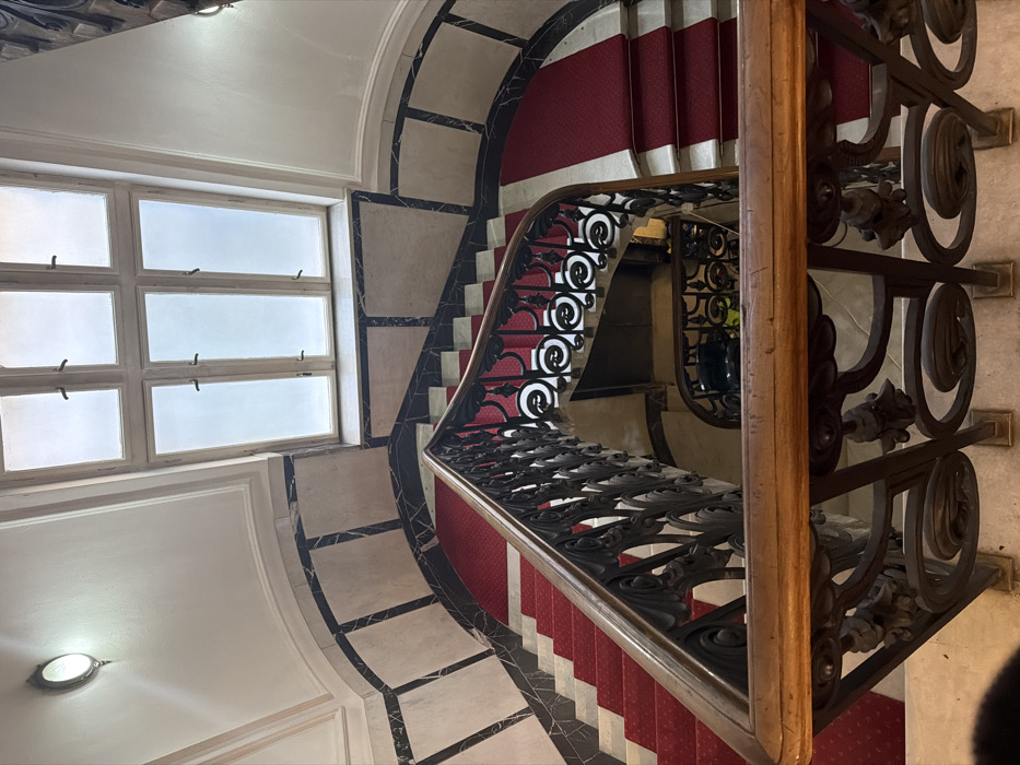 This image depicts the main spiral staircase inside the National Museum of Serbia, located at Trg republike 1a, in Belgrade, Serbia. The scene is observed from a high-angle perspective, looking downward through several turns of the staircase.
The staircase features red carpet runners covering the treads, contrasting with white marble risers and the visible portions of the marble steps. The perimeter of the staircase and landings is bordered with dark, possibly black, marble strips, which frame lighter-colored, square or rectangular marble panels on the adjacent walls. An ornate balustrade crafted from dark metal (likely wrought iron) with intricate scrollwork and decorative elements supports a polished wooden handrail.
At the top of the visible area, a large, multi-pane window, configured as three panes across and two high, allows natural light into the stairwell, displaying an overcast sky. A circular, wall-mounted light fixture with a visible bulb is positioned on the upper left wall, providing additional illumination. No individuals, explicit actions, or interactions are visible within the frame. The overall environment suggests a well-maintained, grand interior space.