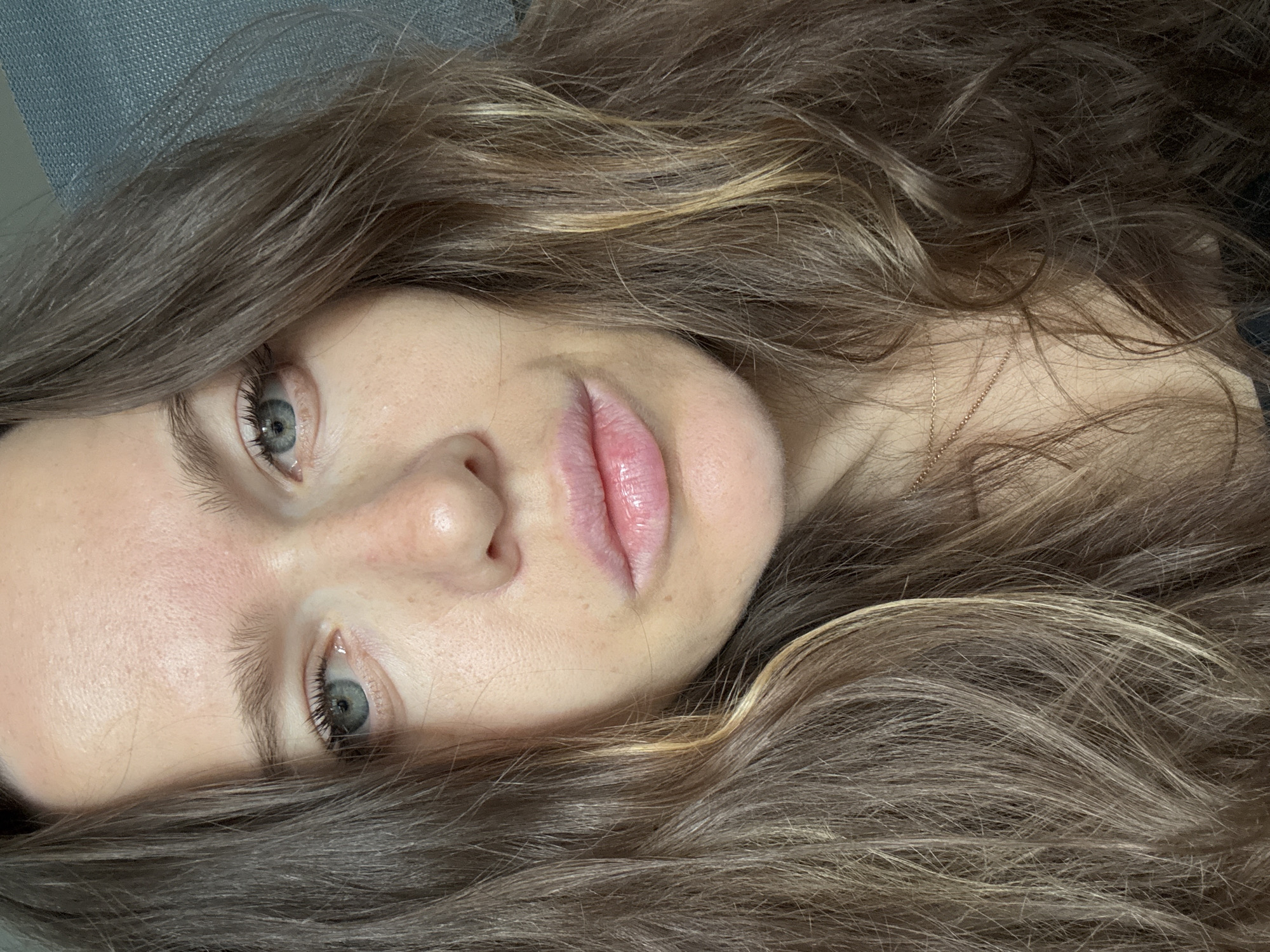 A close-up, angled portrait of a young woman with light skin, gray-blue eyes, and light brown hair with blonde highlights. She is lying down, with her hair spread around her face. Her lips are slightly parted and appear to have a gloss. A thin gold necklace is visible on her neck. The background appears to be a light blue or gray surface. There is no evidence to suggest that the image was taken in the city of Khimki, Russia.