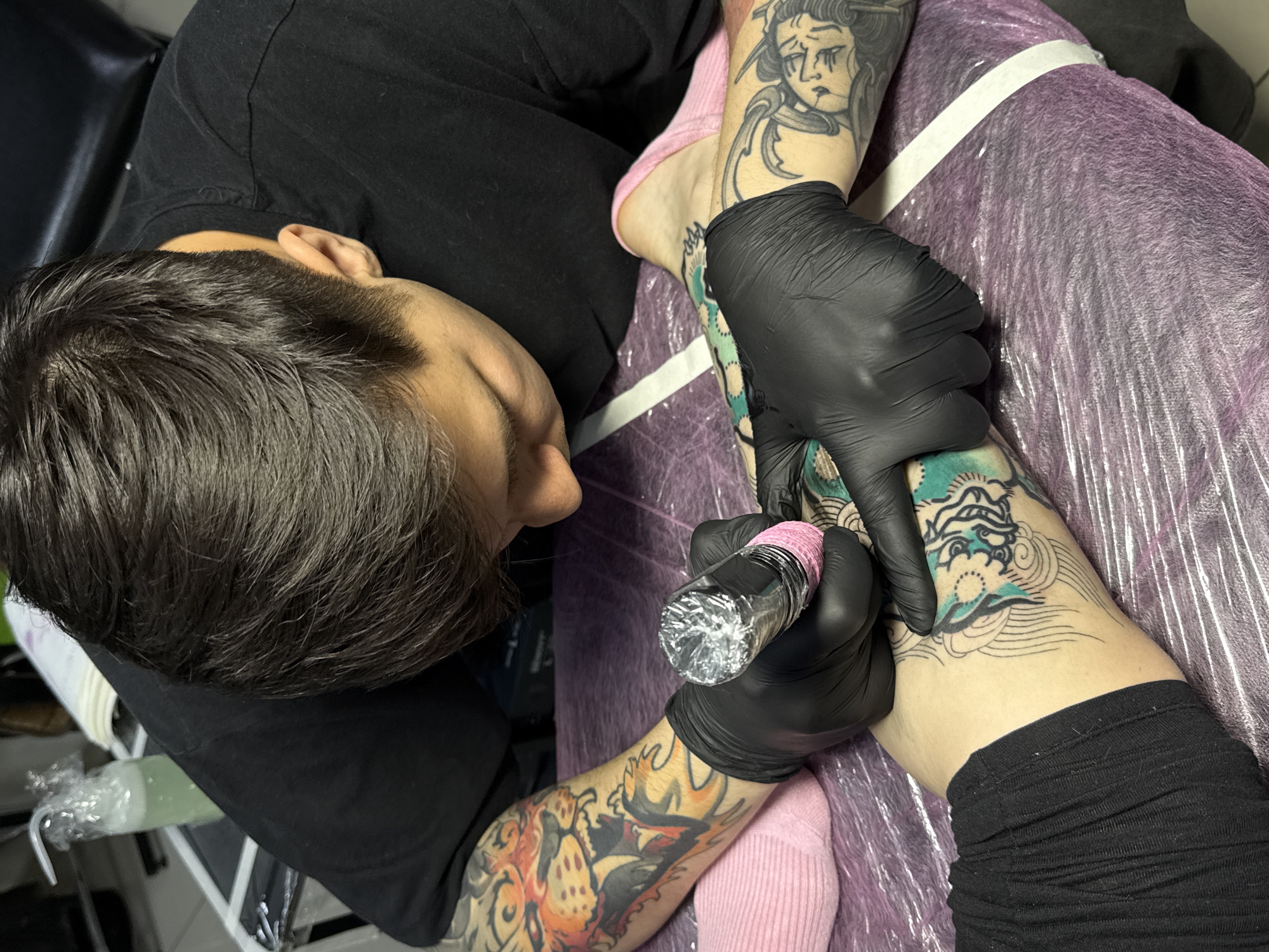 A tattoo artist in black gloves is applying a tattoo to a person's leg. The artist is bent over, focusing on their work. The person receiving the tattoo has existing tattoos on their arms and leg. The procedure is taking place on a surface covered in plastic wrap. There's also a black chair on the left side. The specific location in Rostov-on-Don, Russia cannot be determined from the image.