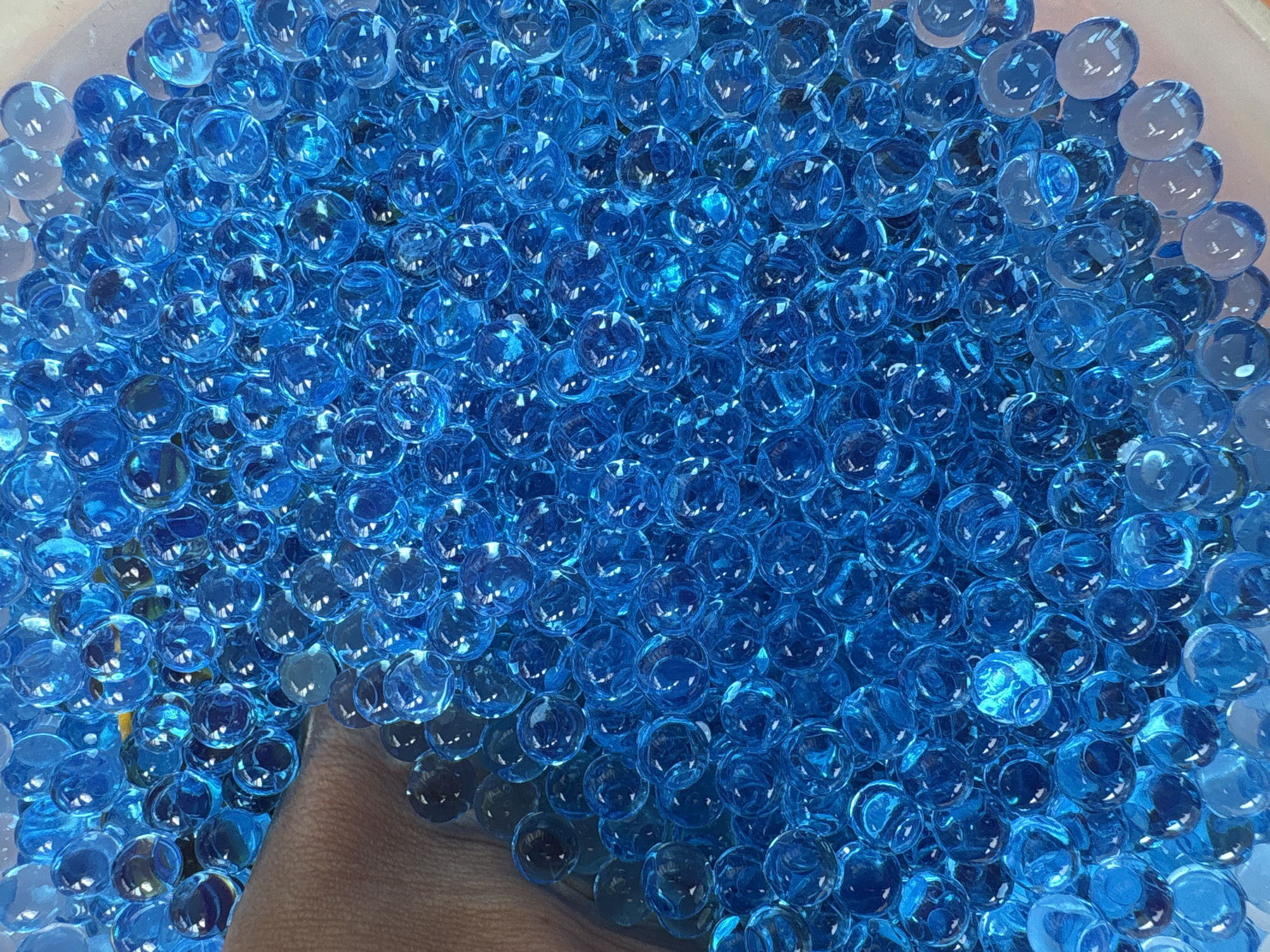 The image is a close-up, overhead shot of a large quantity of blue, translucent orbs, possibly water beads or marbles. A human hand, with skin tone ranging from beige to light brown, is partially visible on the left side of the frame, submerged among the orbs as if reaching into the container. The orbs are tightly packed and fill the majority of the frame. The lighting is bright, causing reflections and highlights on the surfaces of the orbs, giving them a glossy appearance. The background appears to be the inside of a light-colored container, possibly plastic. There is no indication of the city of Ust-Ilimsk, Russia, or any other geographical location in the image.