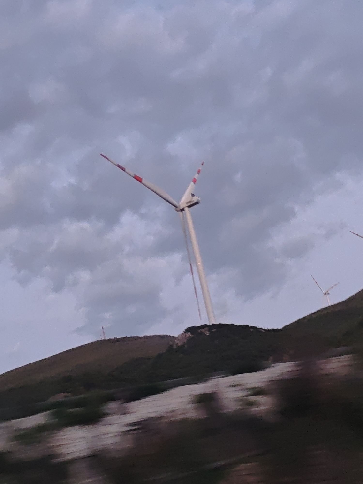 A low-angle shot depicts a wind turbine atop a hill. The turbine is white with red stripes on the blade tips. Another smaller wind turbine is visible in the distance to the right, and a communication tower is present on a nearby hill in the background. The sky is overcast with dark clouds. The foreground is blurry, suggesting the image was taken from a moving vehicle. The location is Δημοτική Ενότητα Προμαχώνος, Greece.