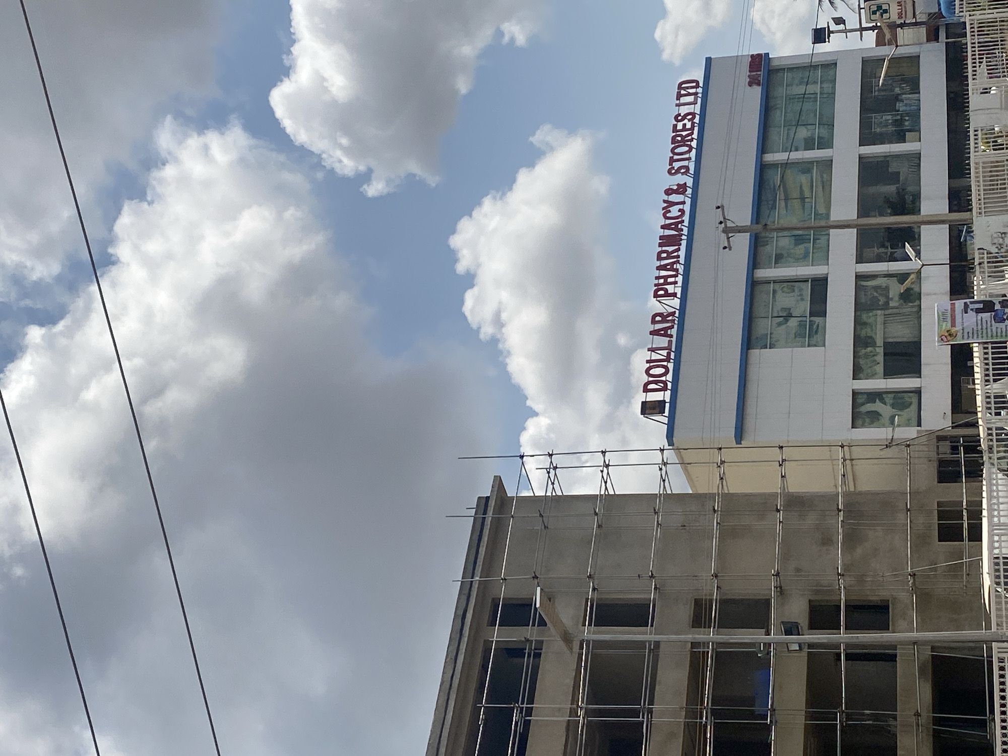 This is a slightly low-angle, full-shot, eye-level, outdoor, daylight, medium-distance photograph of a building under construction next to a pharmacy in Ado, Nigeria. The building on the left is a concrete structure with exposed rebar and scaffolding covering its exterior, indicating ongoing construction. The building on the right is a white, multi-story commercial building with a blue trim and red lettering that reads "DOLLAR PHARMACY & STORES LTD." The pharmacy sign also indicates "24 HRS." The sky is partly cloudy with large white cumulus clouds against a light blue sky. There are two dark wires running horizontally across the upper portion of the image. A utility pole is visible in front of the pharmacy. The lower part of the image shows a white metal fence and the top of a white vehicle.