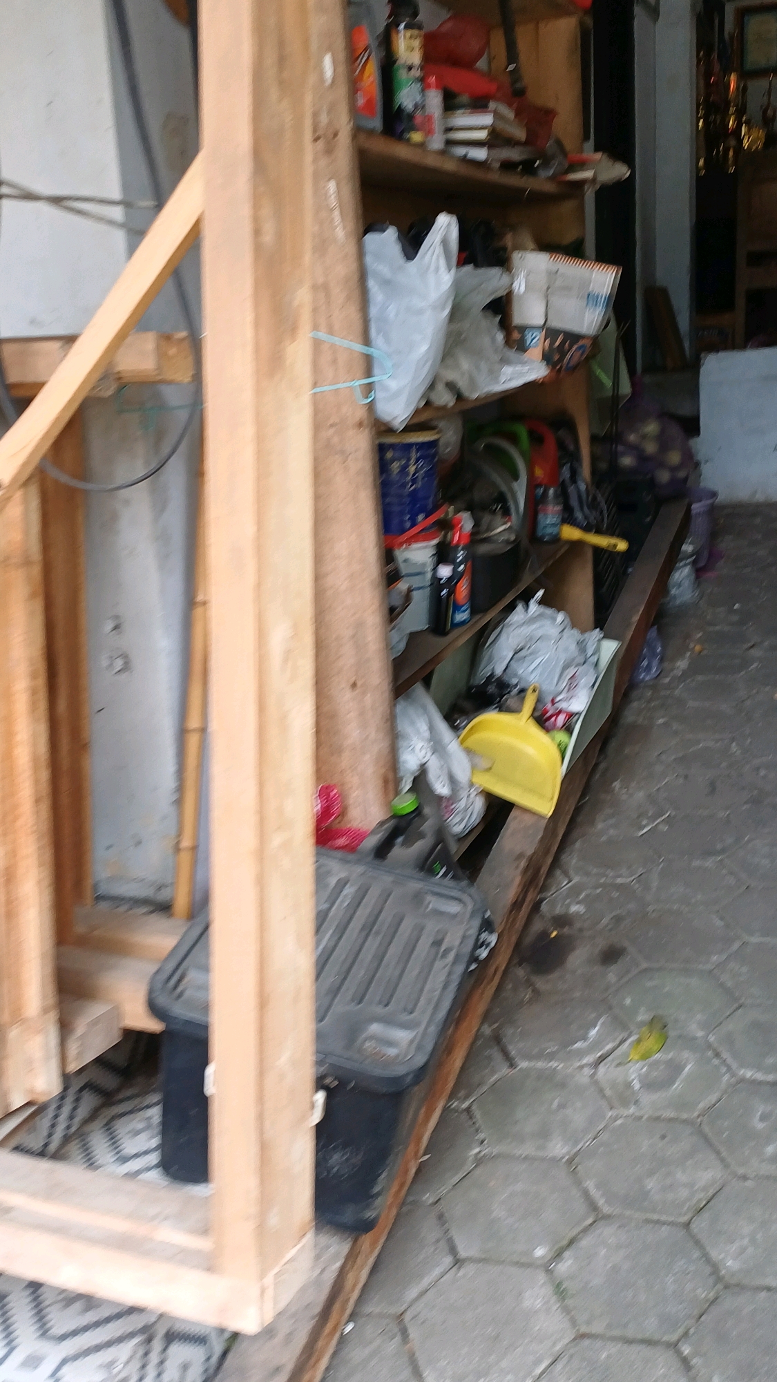 The image depicts an outdoor storage area adjacent to a building in Karangwuni, Indonesia. A wooden frame structure is visible on the left, part of what appears to be a doorway or entrance. To the right, a set of wooden shelves is filled with various items, including plastic bags, bottles, a blue bucket, and what looks like cleaning supplies. A yellow dustpan is placed on the lower shelf. On the ground in front of the shelves, there is a black plastic storage container. The ground is paved with hexagonal concrete tiles. The scene is cluttered and appears to be a functional, albeit disorganized, storage space. There are no people visible in the image.